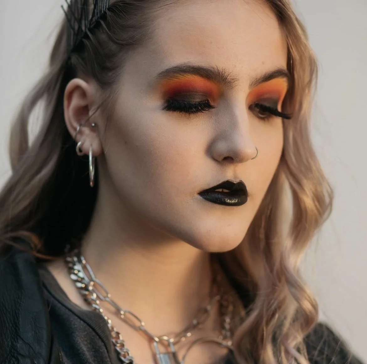 Close-up of a woman with bold makeup, including orange eyeshadow, black lipstick, and glittery black eyeliner. She has earrings, layered necklaces, and wavy blonde hair.