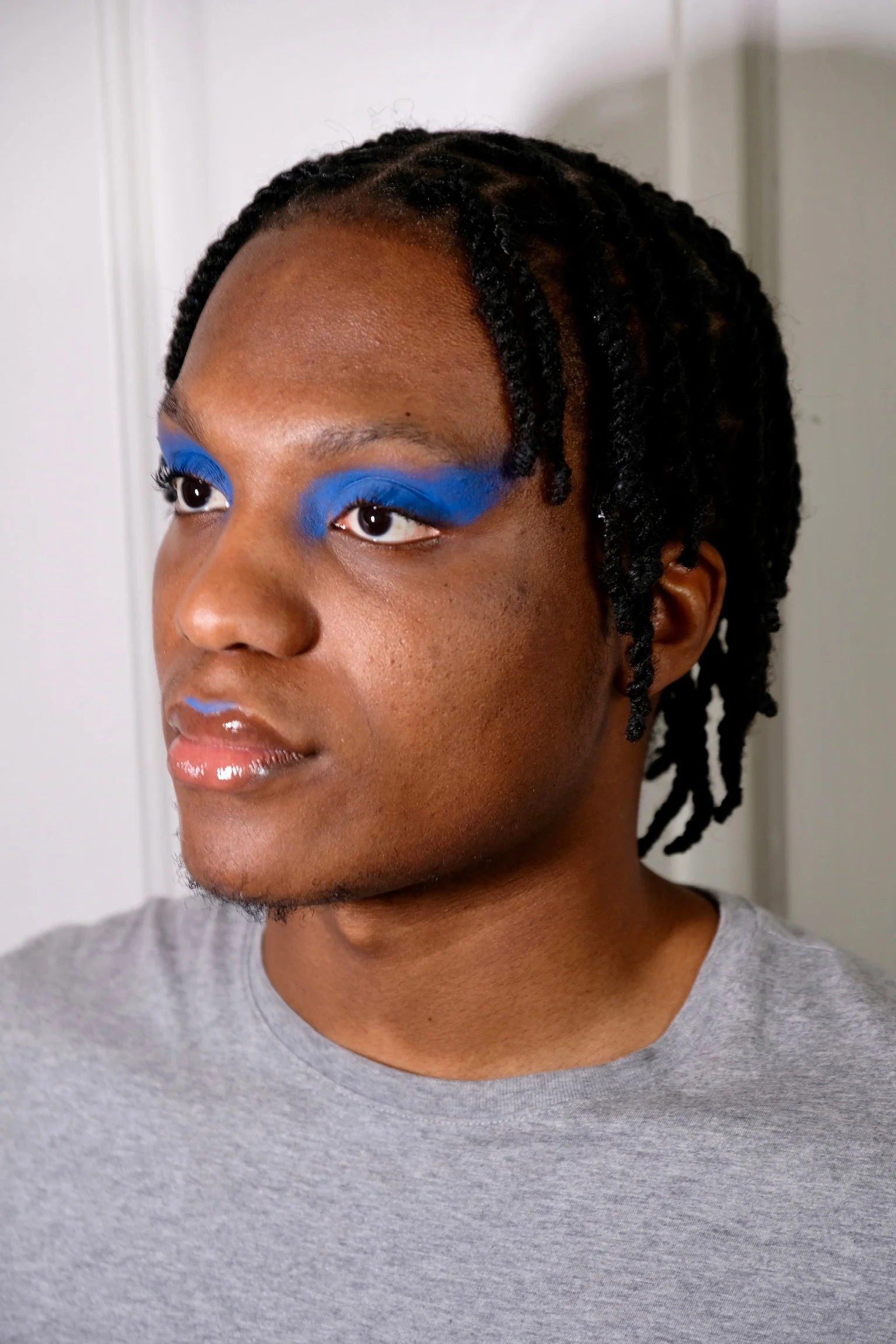 Close-up of a person with dark skin and dreadlocks styled with twists, wearing bright blue eyeshadow, a shiny lip gloss, and a grey t-shirt, looking slightly to the side.