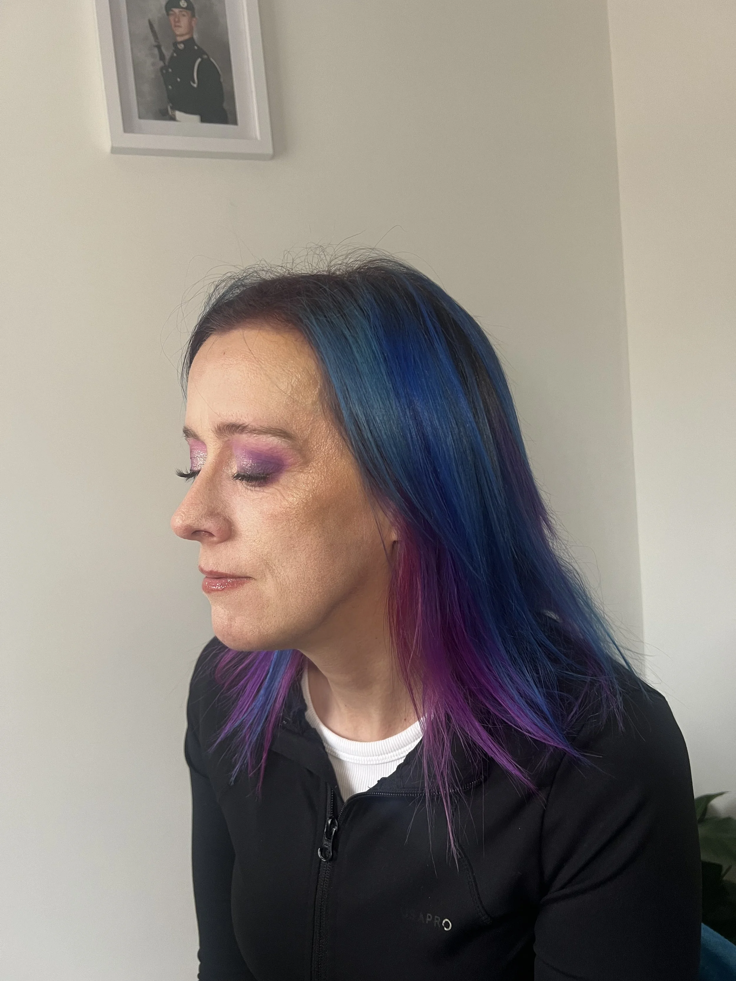 A woman with vibrant, rainbow-colored hair, featuring shades of purple, blue, and pink, is shown in profile with her eyes closed and wearing eyeshadow, against a plain light-colored wall. A framed photograph of a young man in a military uniform is ha