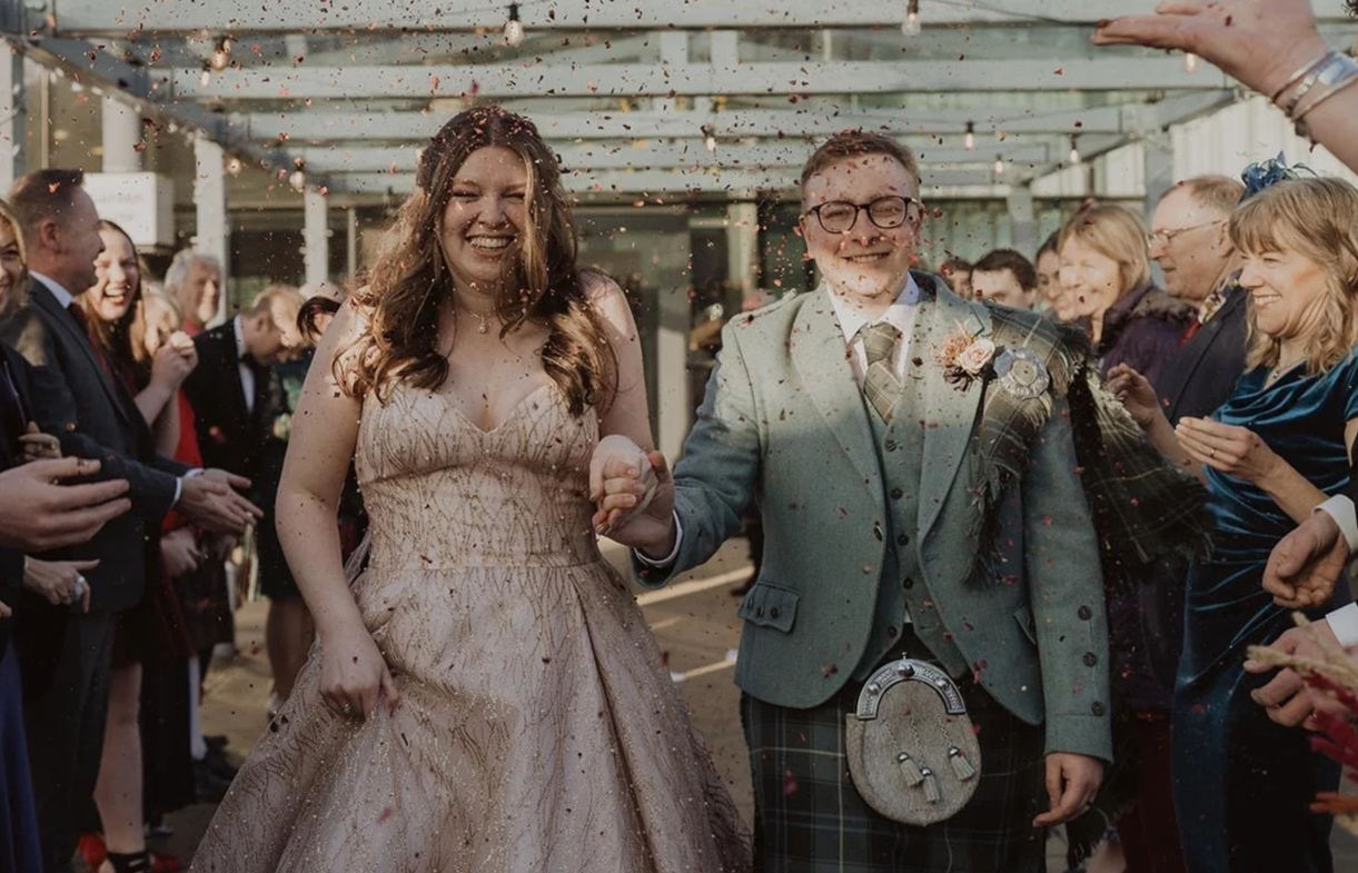 A bride and groom walking hand in hand through a crowd, celebrating a wedding with confetti and happy guests.