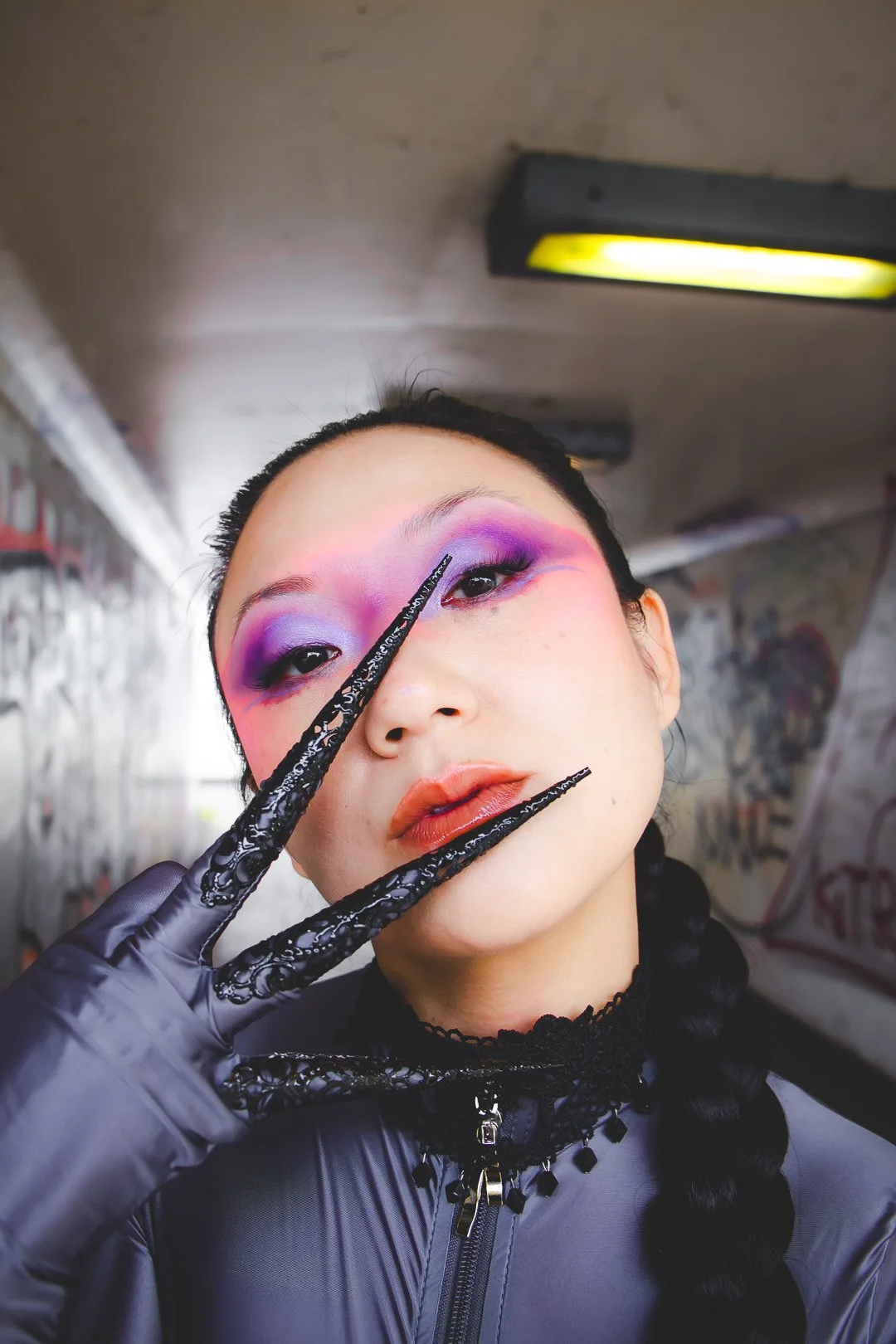 Person with dramatic purple and pink eyeshadow, wearing black gloves, holding a black lace knife, in an urban graffiti-covered setting.