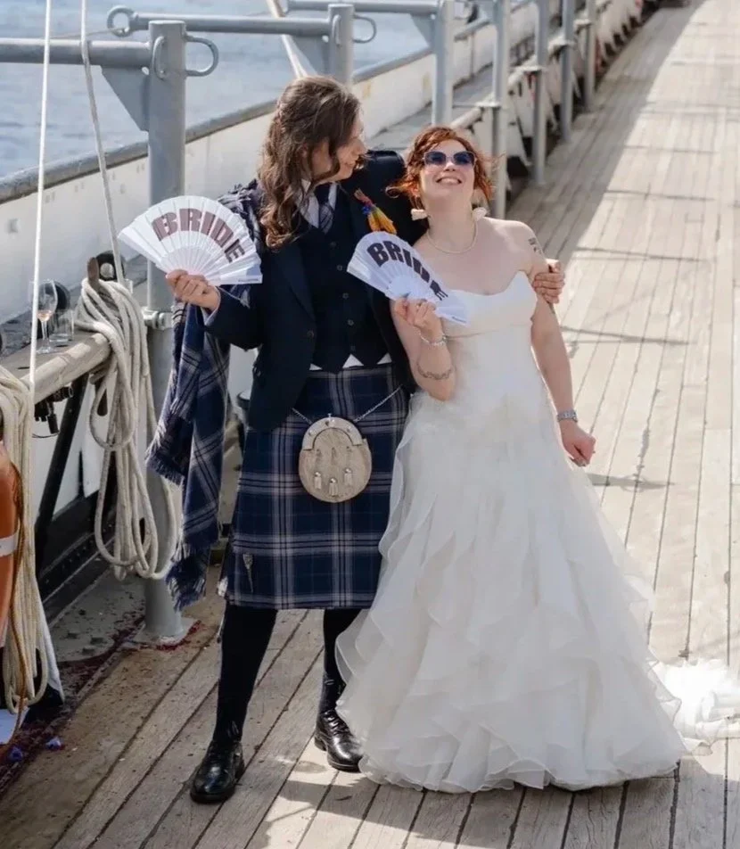 Two women, one in a wedding dress and the other in a tartan kilt, sharing a joyful moment on a boat dock with a wooden walkway and water beside them. They are holding fans that say "BRIDE".