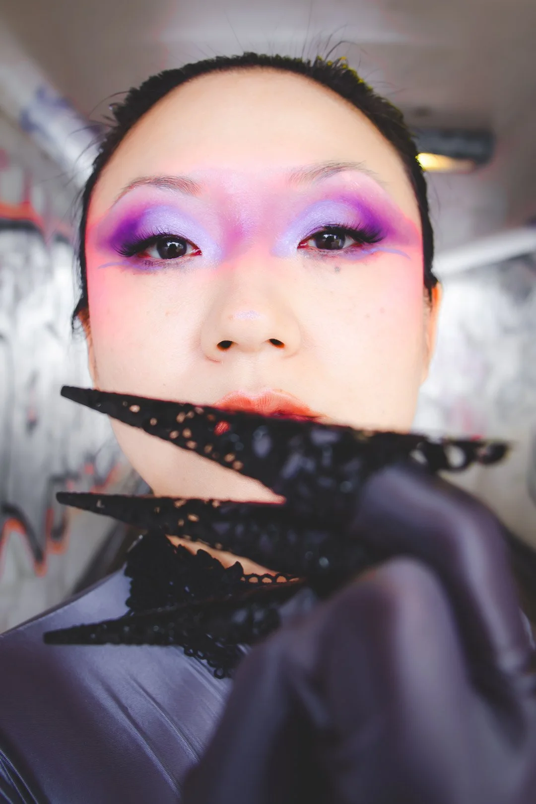 Close-up of a person with colorful purple and pink makeup on their eyes, wearing black lacy gloves, and holding an ornate black hand accessory near their face.