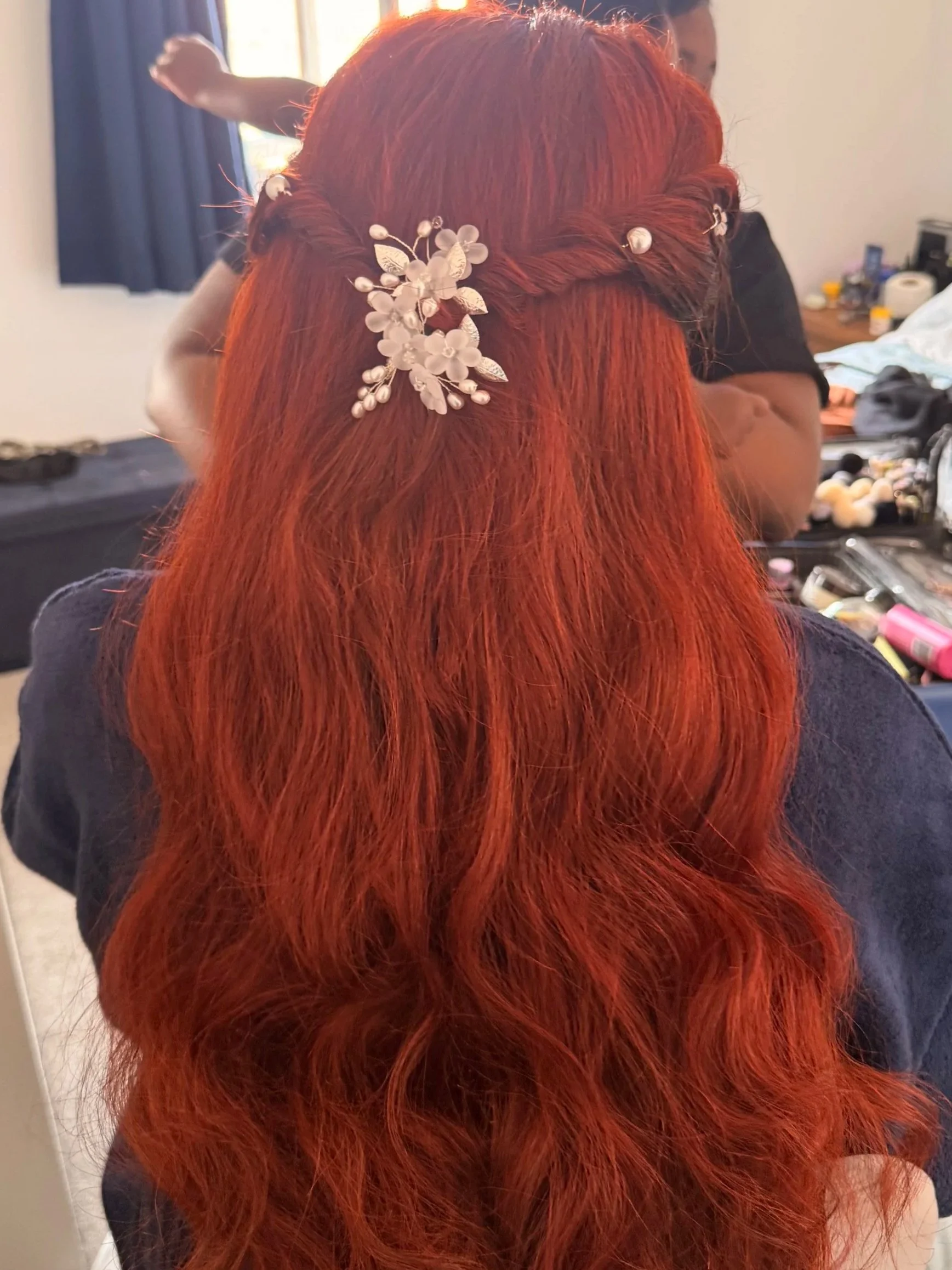 A woman with long red hair styled in a half-up, half-down braid decorated with white floral hair accessories, standing in a room with makeup supplies on a table.