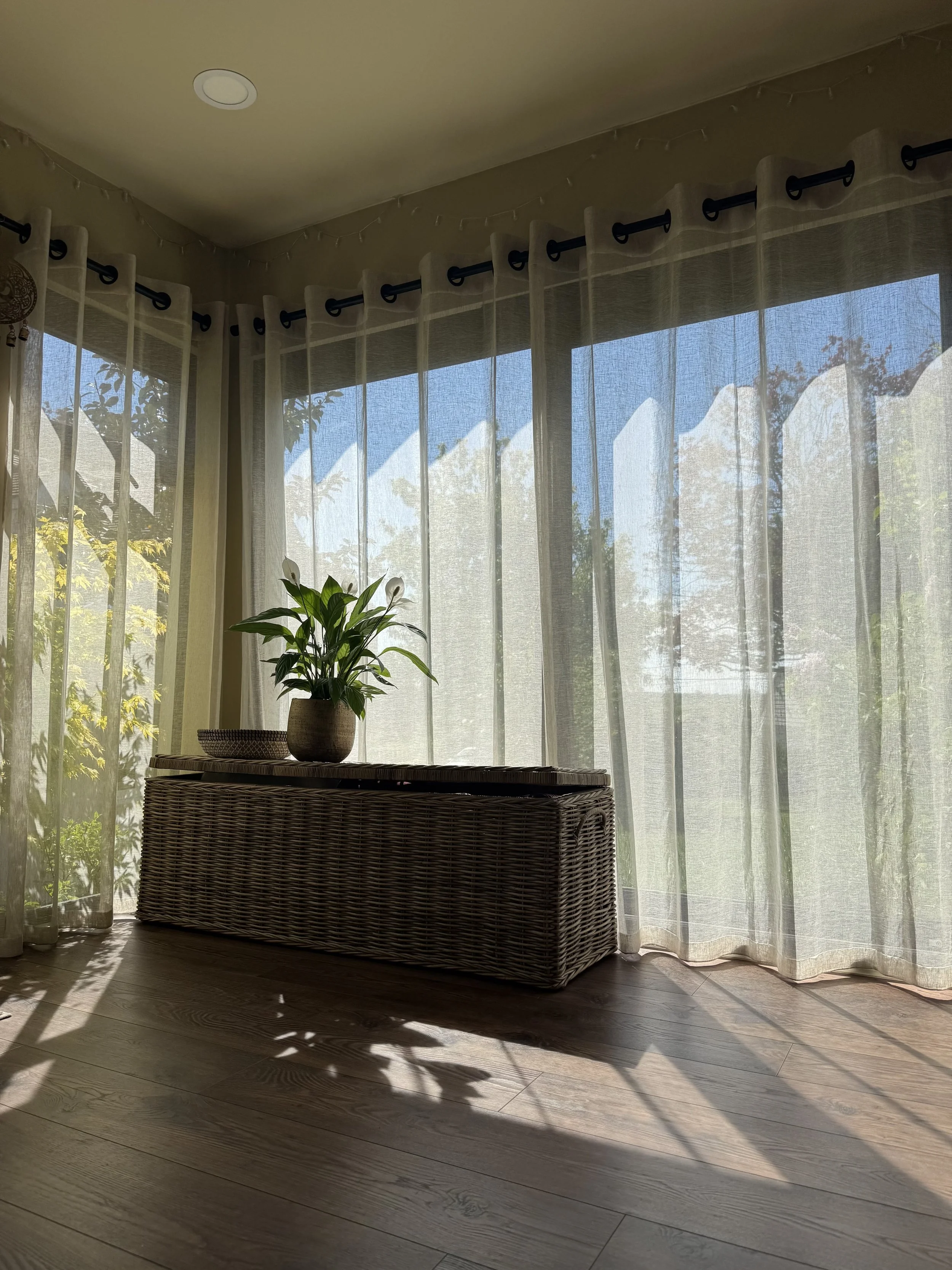 Gentle sunlight streams into truly.madly.yoga home yoga studio, casting shadows on a wooden floor near a wicker table with a potted plant and a woven basket. Sheer curtains cover a large window, with greenery visible outside.