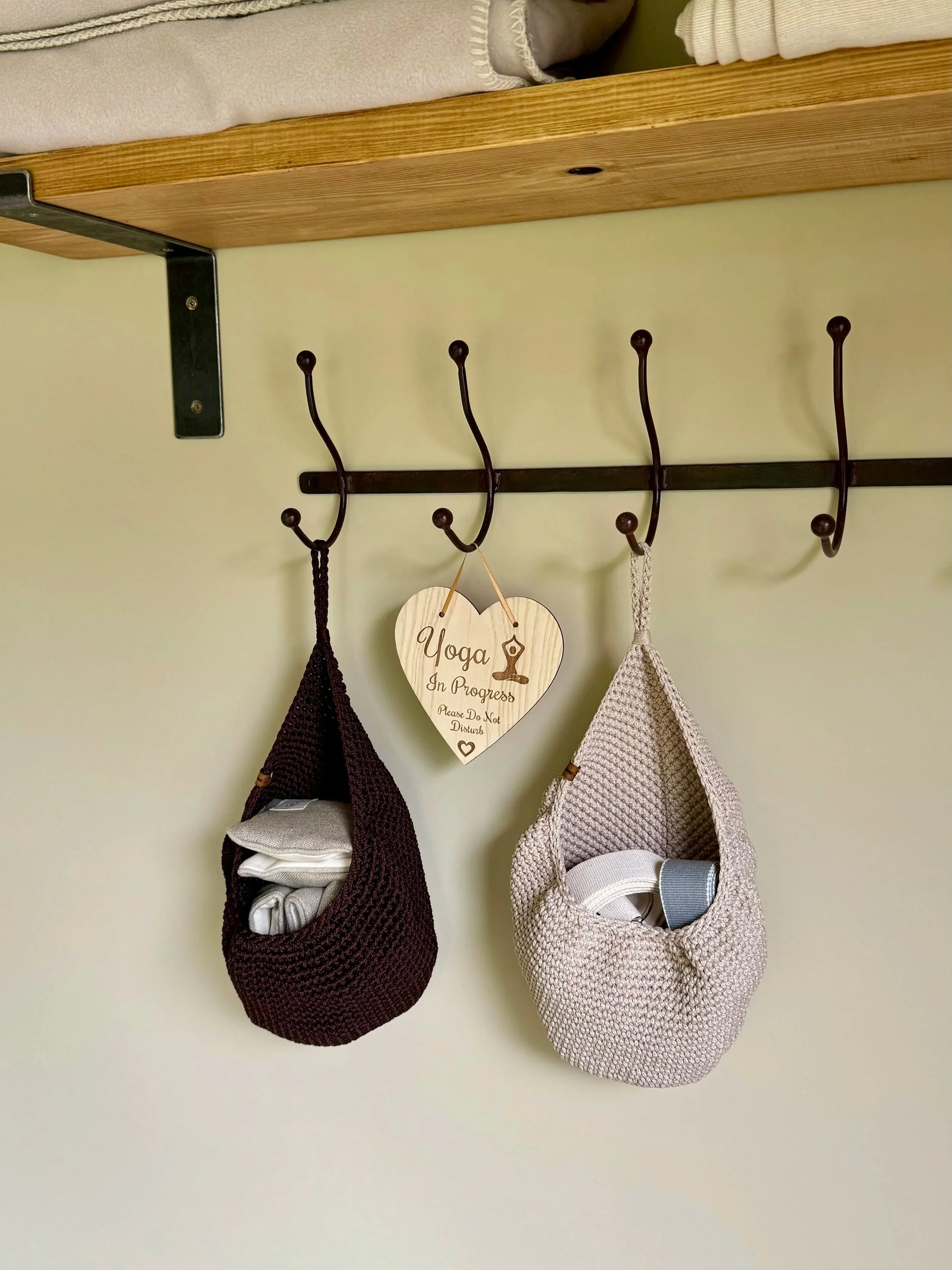 Two hanging stuffed bags, one dark brown and one beige, on a wall-mounted hook rack. A wooden sign that says "Yoga in Progress, Please Do Not Disturb" hangs between them in truly.madly.yoga home yoga studio.