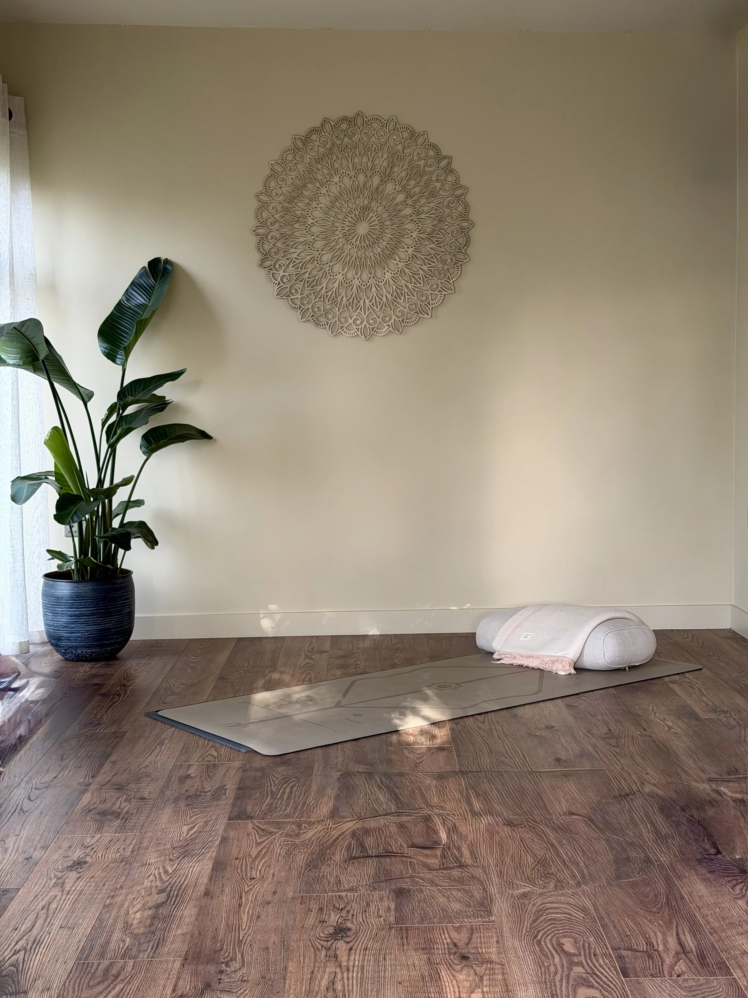 truly.madly.yoga home yoga studio with hardwood flooring, including a yoga mat, a bolstered cushion with a blanket, and a potted plant near a window with sheer curtains, a decorative wall hanging, and sunlight streaming in.