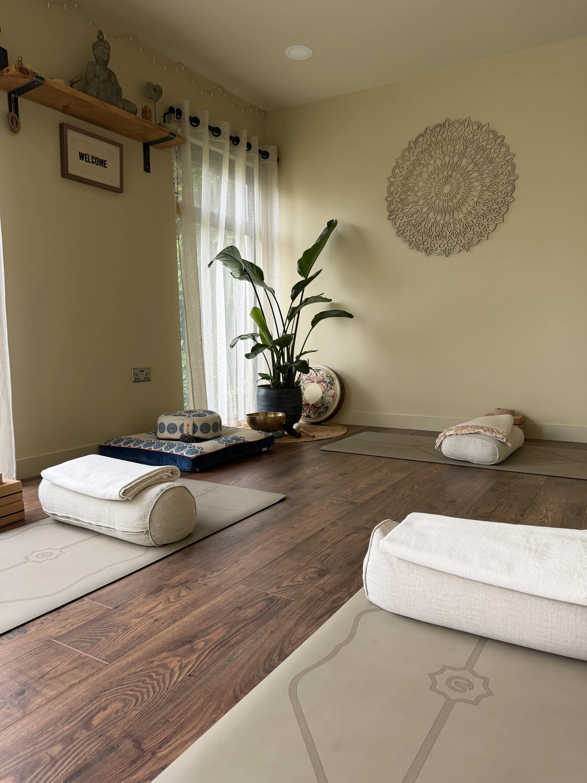 truly.madly.yoga tranquil yoga room with mats, bolsters, and a large green plant near a window with sheer curtains, decorated with wall art and a wooden shelf with decorative objects.