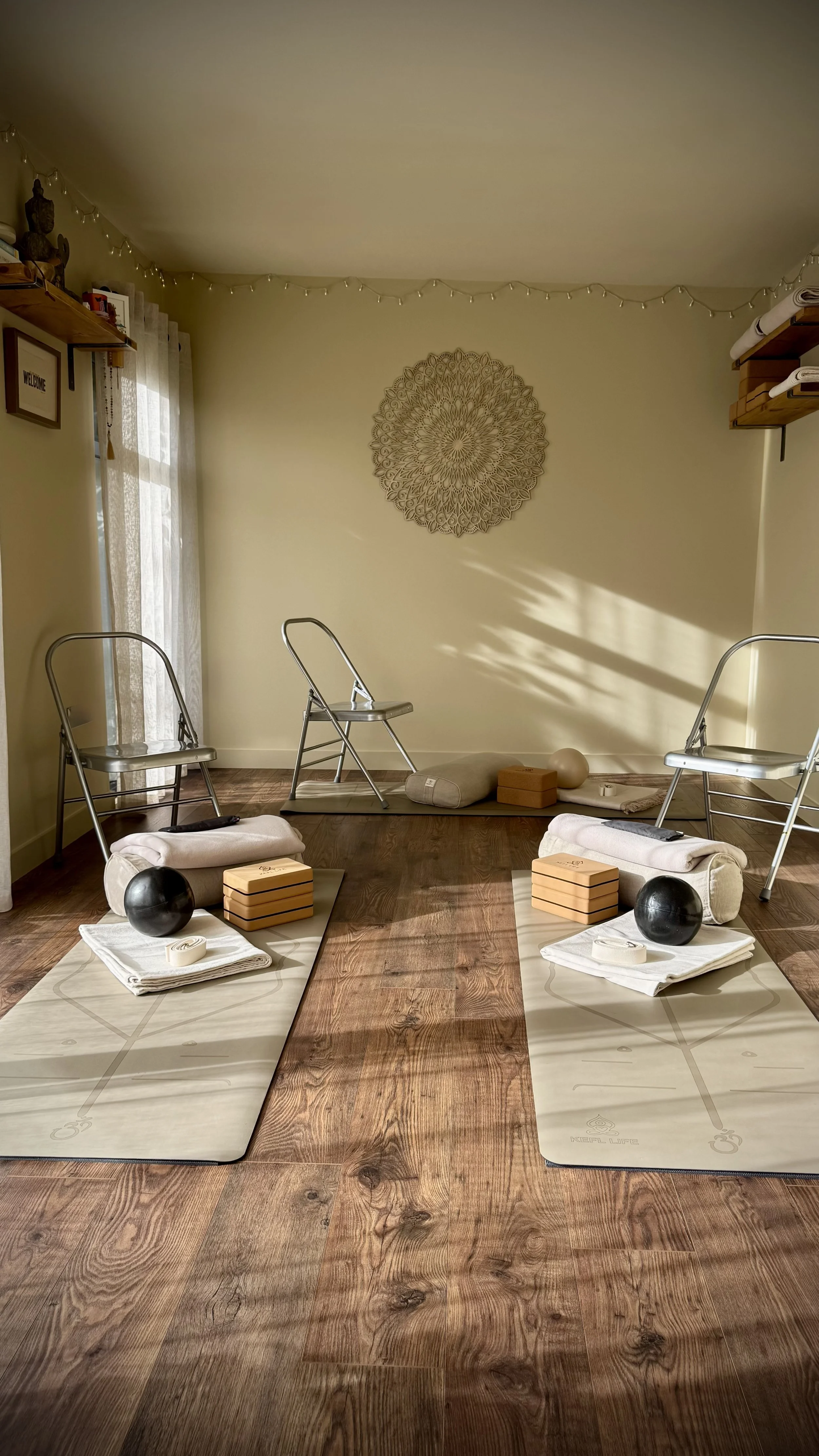 truly.madly.yoga yoga studio with two mats laid out on a wooden floor, each with a rolled towel, blocks, and a ball. Three metal chairs are positioned behind the mats, and a decorative hanging is on the wall.
