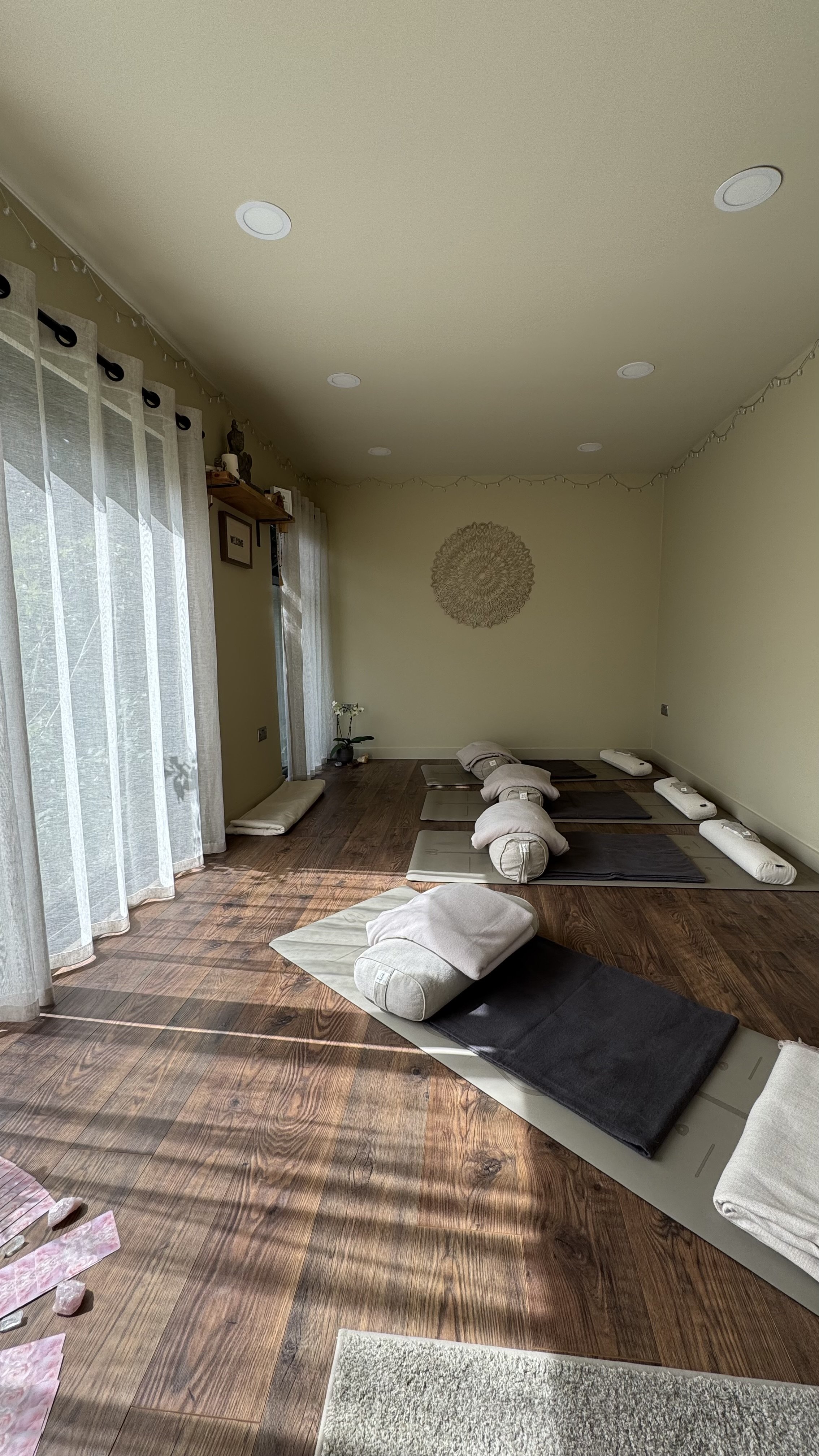 truly.madly.yoga home yoga studio with mats, bolsters, and blankets arranged on a wooden floor, natural light coming through sheer curtains.