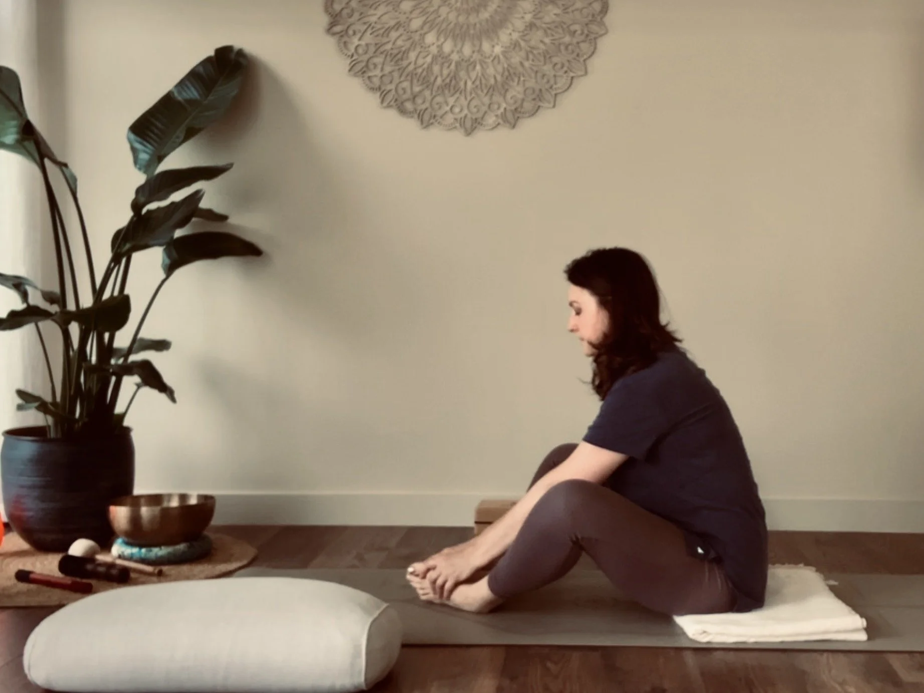 truly.madly.yoga sitting on a yoga mat, stretching her legs and reaching towards her toes in her cosy at home yoga studio with a large potted plant and decorative wall art.