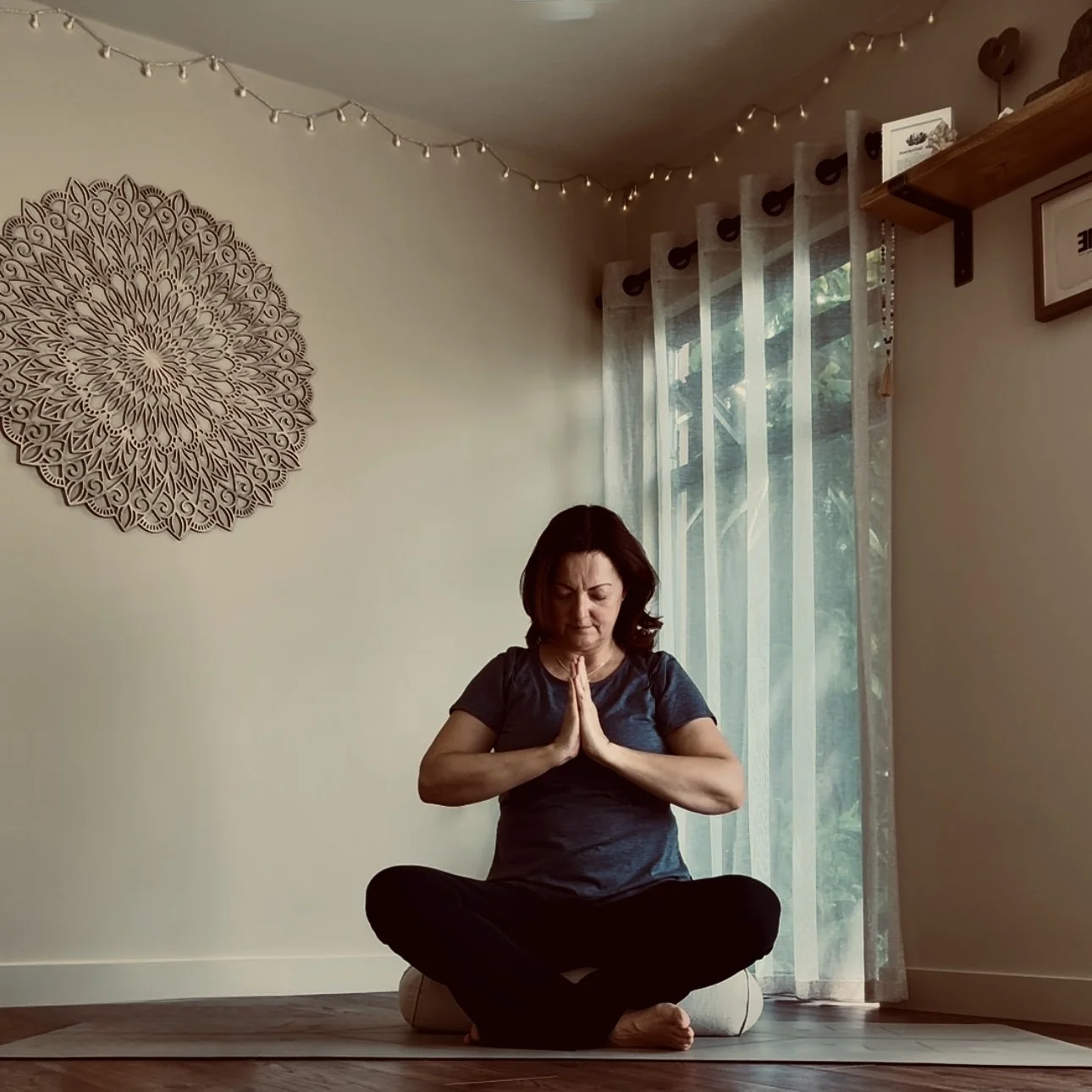 truly.madly.yoga practicing mindfulness meditation in her peaceful home yoga studio with decorative wall art, string lights, and sheer curtains.
