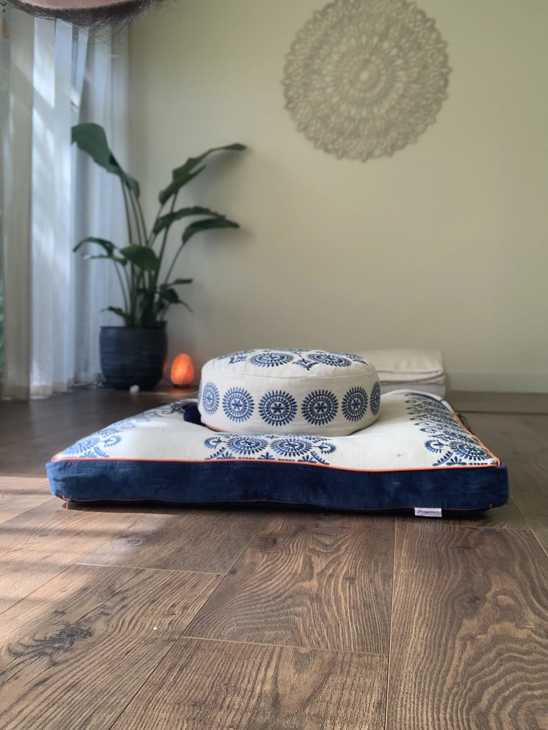 truly.madly.yoga teaching cushion with blue eastern design on a wooden floor, with a plant, wall decoration, and a salt lamp in the background of her home yoga studio.