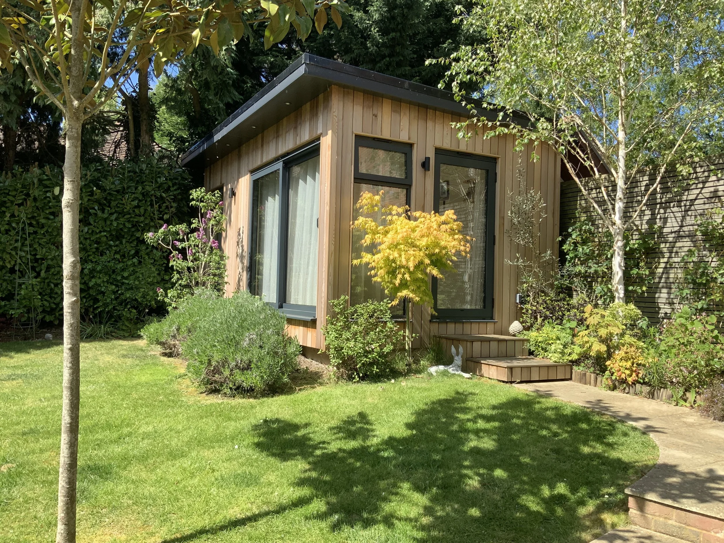 truly.madly.yoga cosy home yoga studio, a small but airy modern wooden studio, surrounded by green trees and plants in a garden with a stone pathway, under a bright blue sky.
