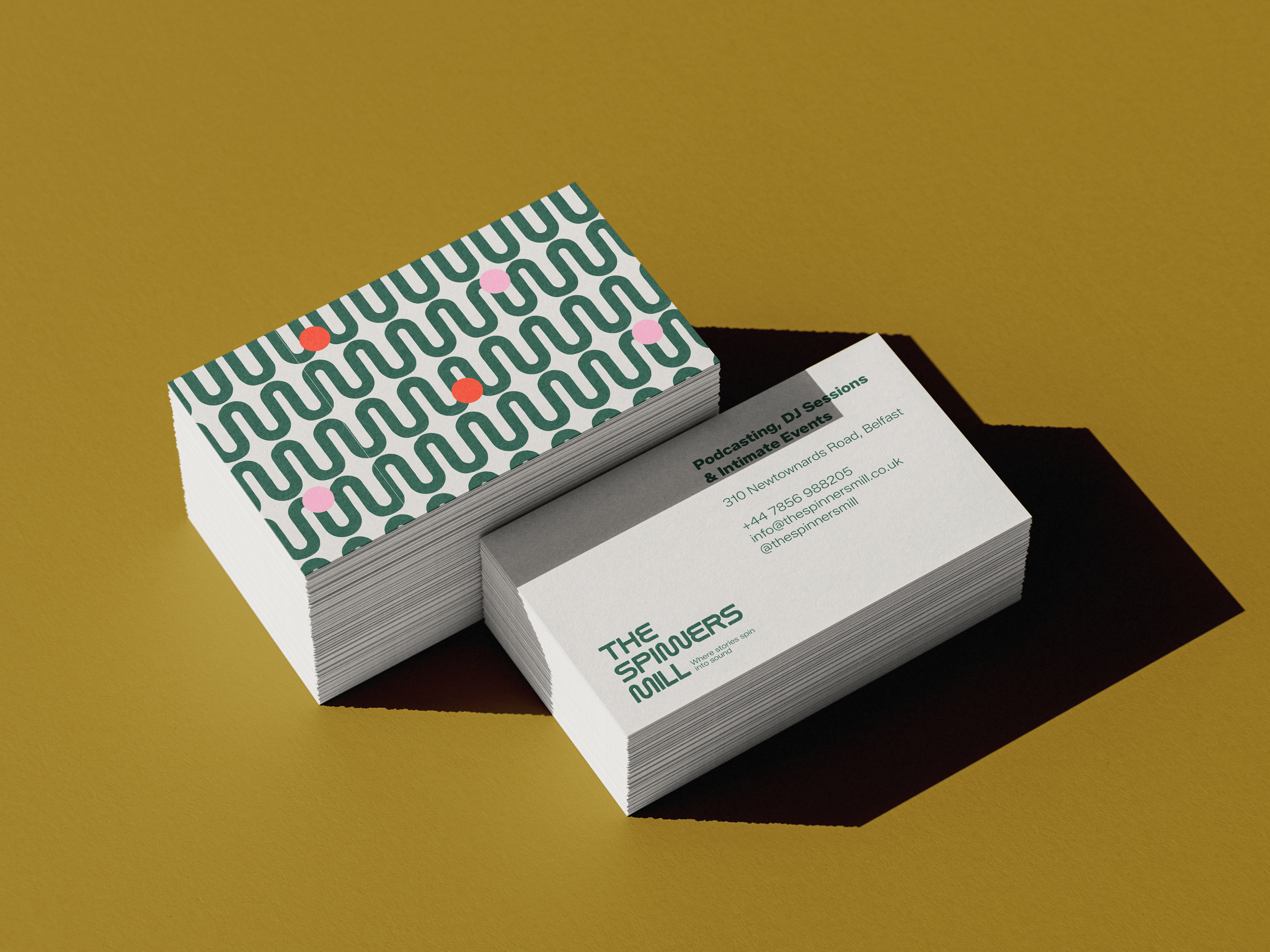 A stack of business cards with green and pink geometric pattern on top, placed on a larger paper with contact information for The Spinners Mill event organizers, against a yellow background.