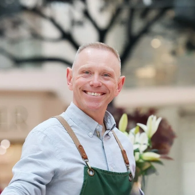 A smiling man wearing a light blue shirt with a green apron, standing outdoors with blurred background trees and flowers.