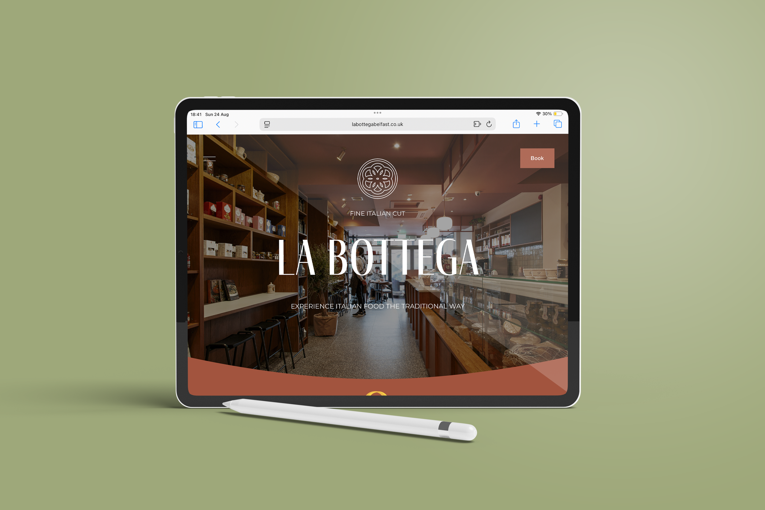 An iPad with a stylus displaying a restaurant website called La Bottega, showing an interior view of a cozy Italian eatery with shelves, seating, and a counter, against a soft green background.