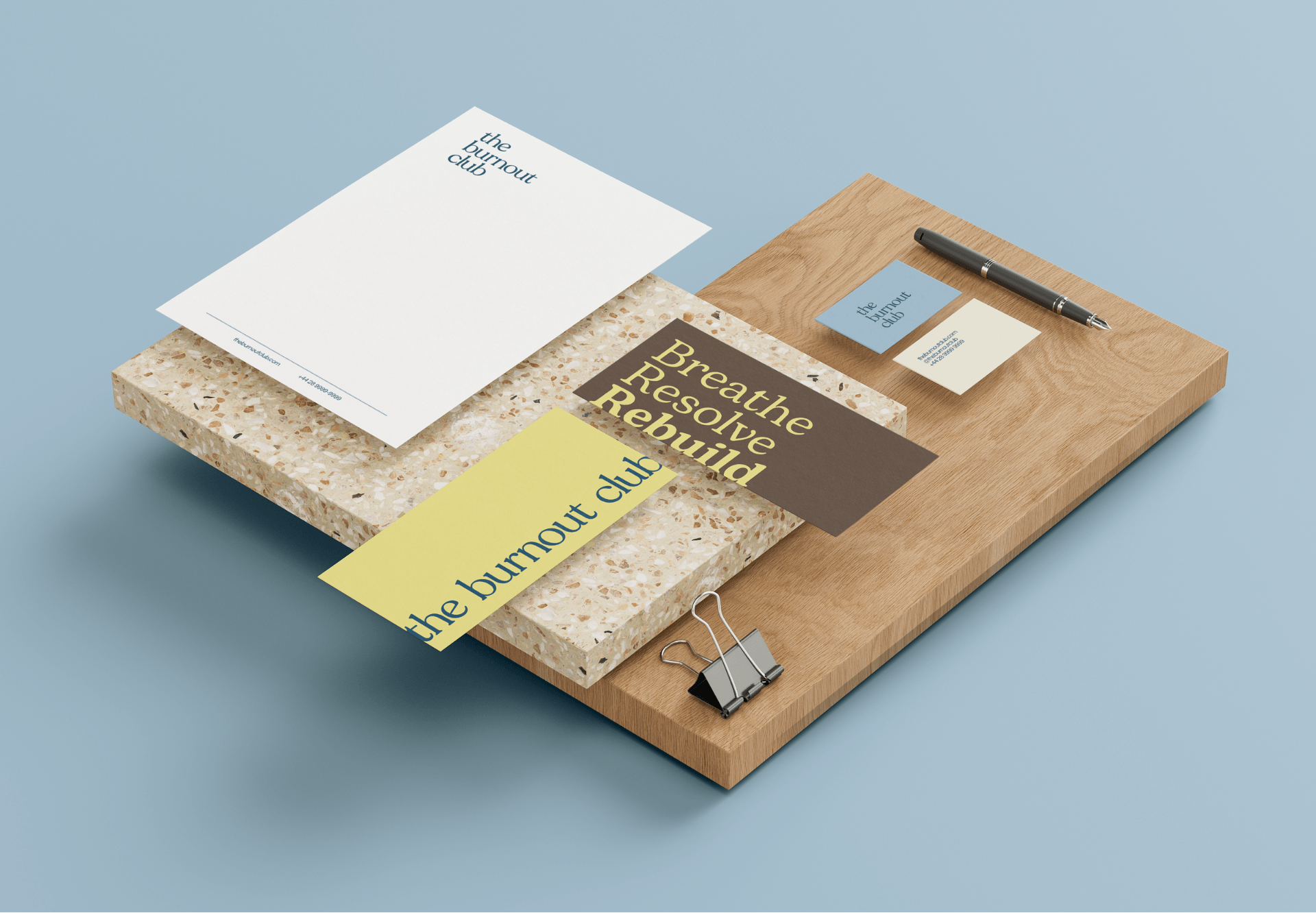 Business stationery and branding materials on a wooden desk, including business cards, flyers, and a pen with the company name 'the burnout club'.