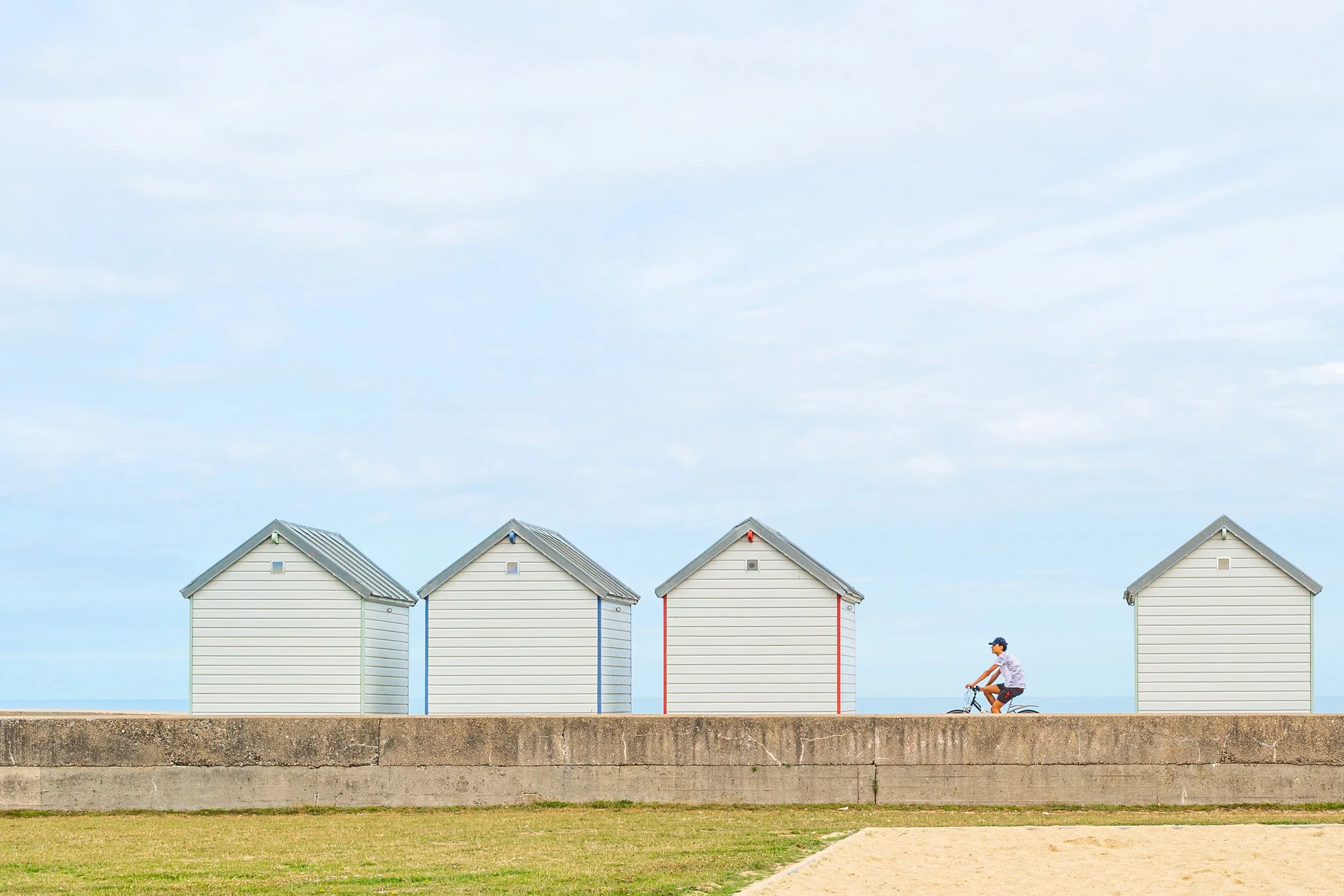 Vier pastelgekleurde strandhuisjes staan uitgelijnd langs de waterkant onder een bewolkte blauwe lucht, terwijl een persoon op een fiets voorbijrijdt.
