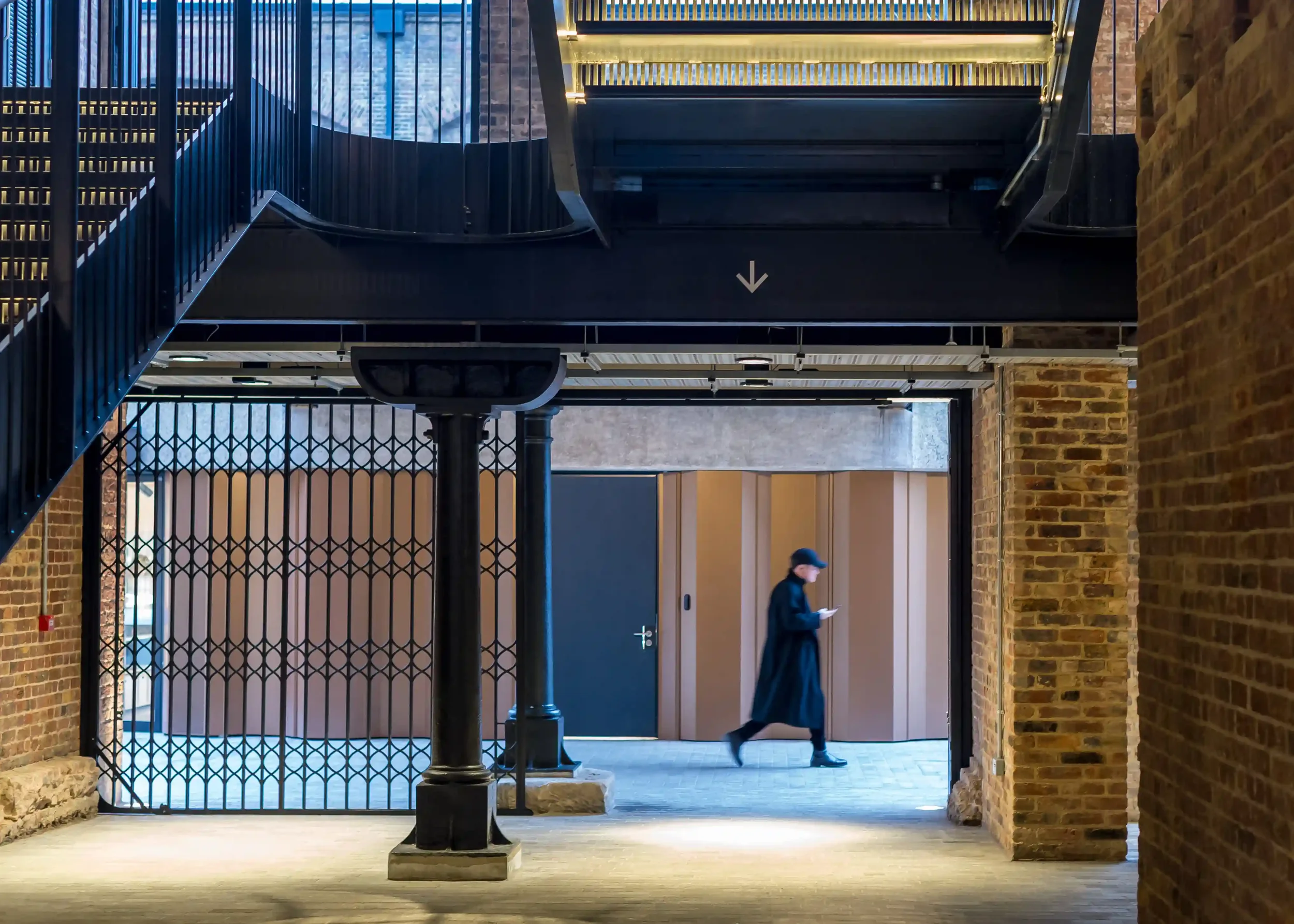 Een persoon loopt door een gebouw met bakstenen muren en metalen trappen, onder een zwarte sprinkler en een pijl die naar beneden wijst. Pakhuis in Londen. Lijnen spel van openingen met warme tinten, 