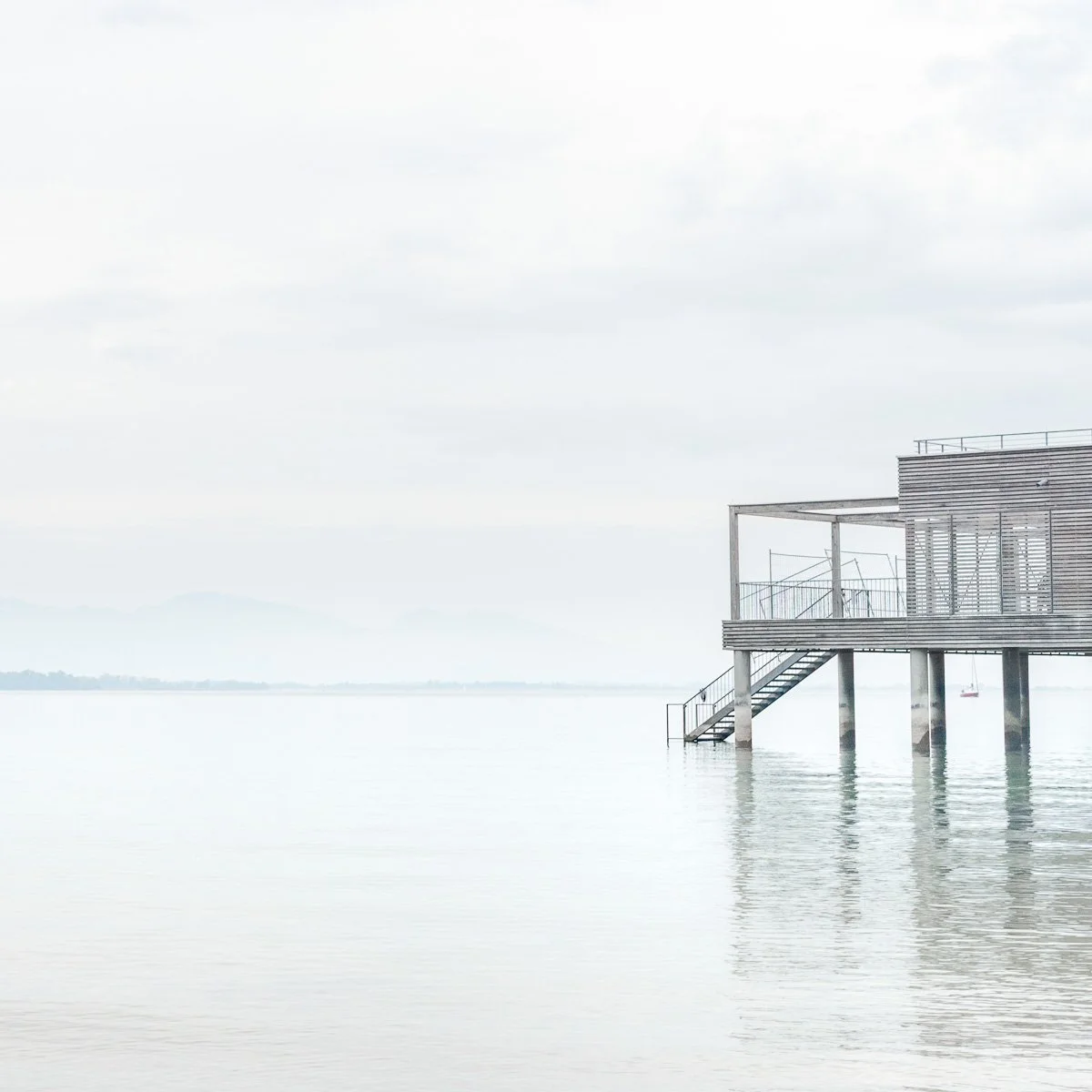 Een modern houten gebouw op palen boven het water, met een trap die naar het water leidt, onder een bewolkte lucht. De Bodensee met de zwitserdse bergen op de achtergrond. Verstild beeld met veel lichte tinten. 