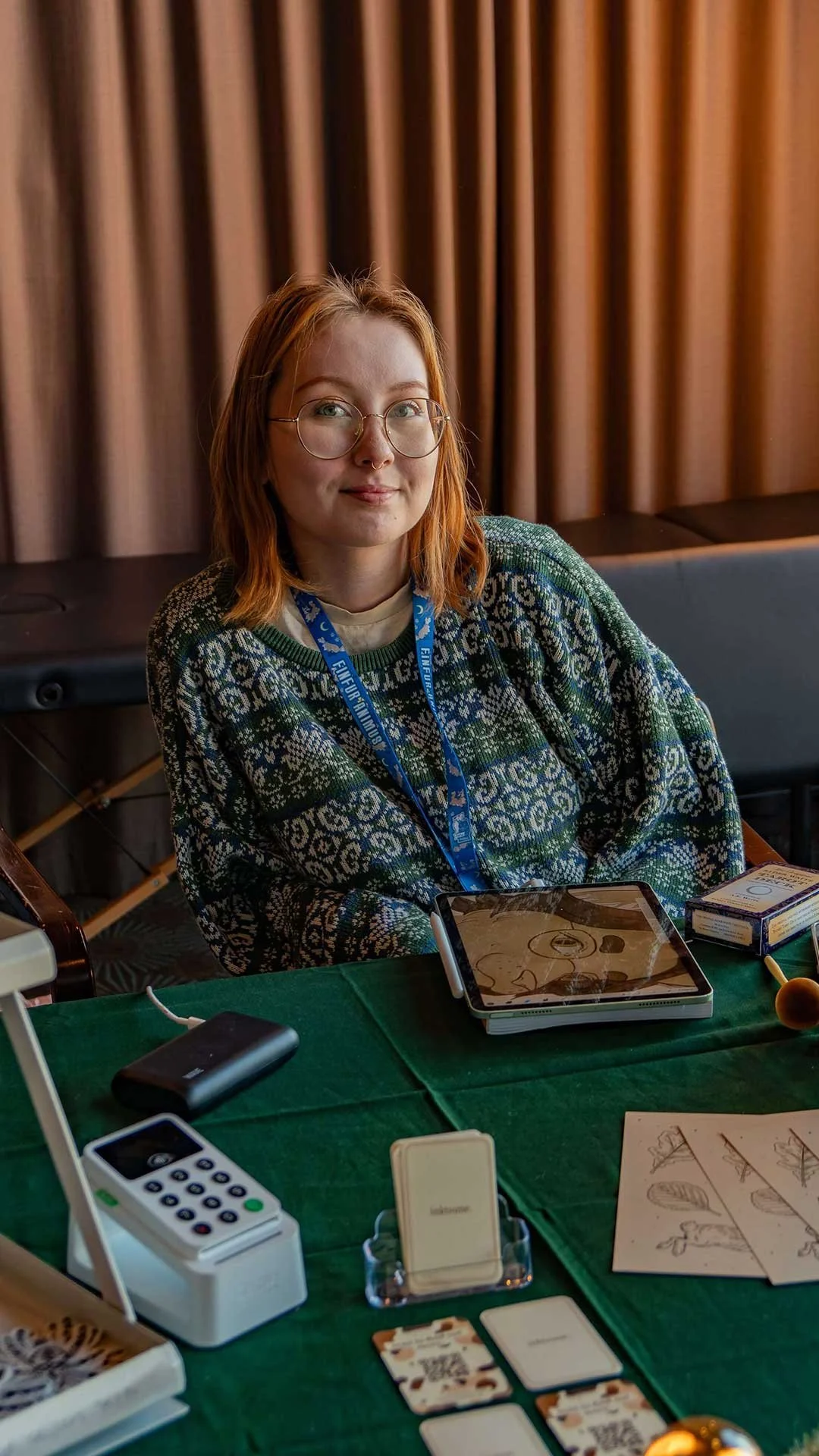 A young woman with glasses and red hair sitting at a table covered with a green cloth, various electronic devices and cards, and a box of cigarettes, with brown curtains in the background.