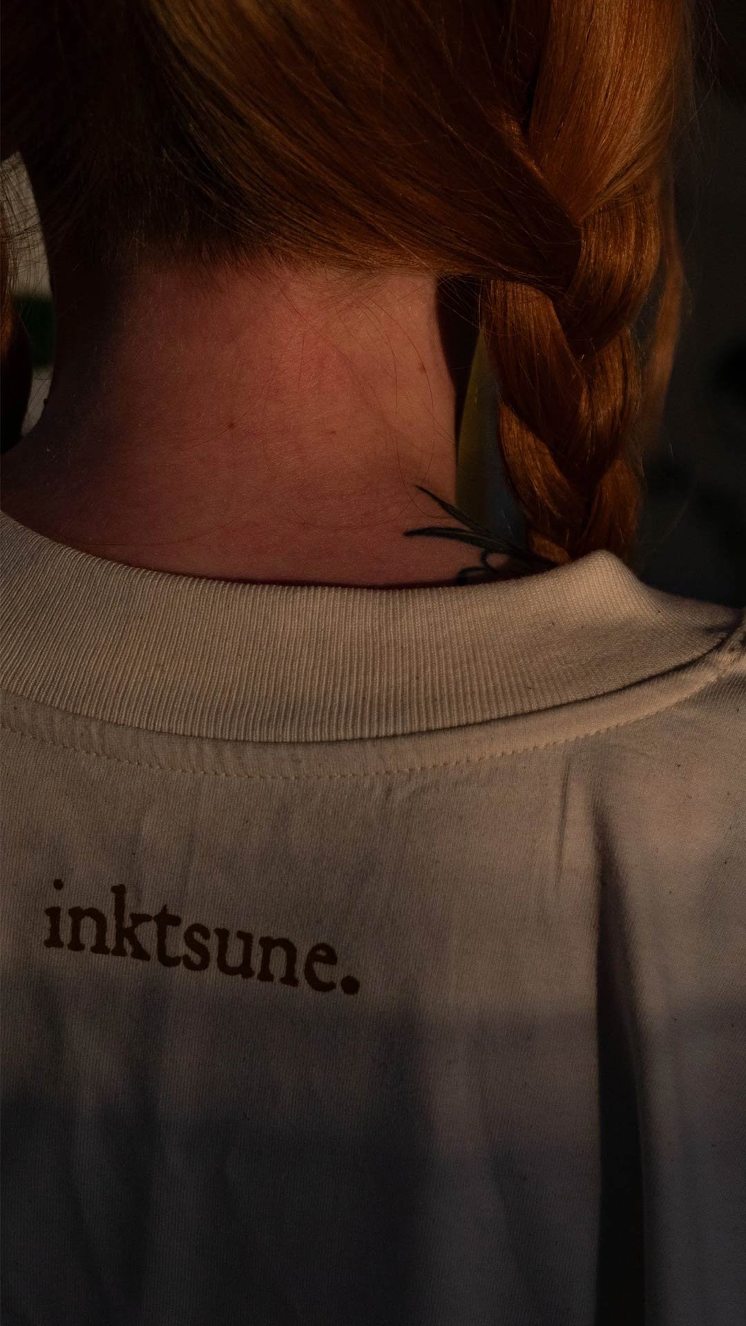 Back of a person with red hair in a braid, wearing a beige hoodie with the word 'inktsune.' printed on it.