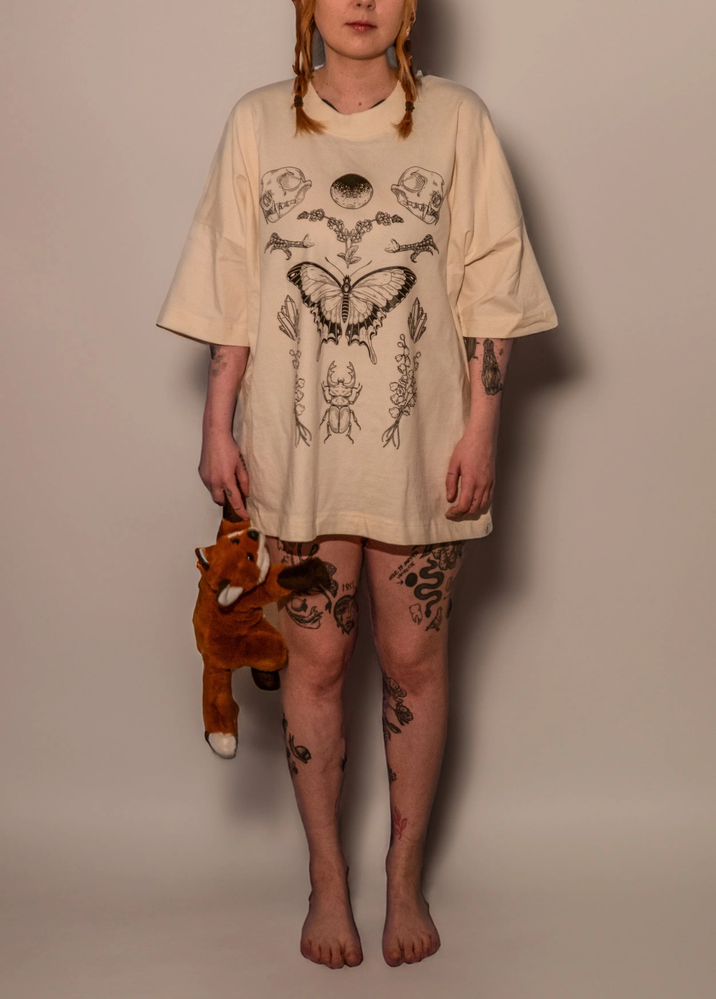 A woman with red hair styled in two braids, wearing an oversized beige t-shirt with black line art of various insects, skulls, and a butterfly on the front. She is barefoot, standing against a plain background, and holding a plush fox toy in her left hand. She has multiple tattoos on her legs and arms.
