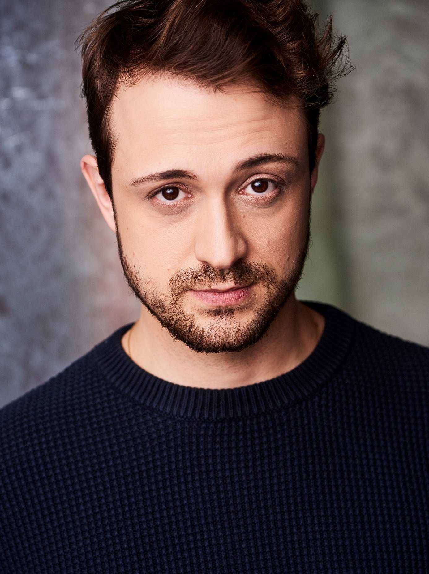 Close-up of a young man with short dark hair, brown eyes, and a beard, wearing a dark textured sweater, against a blurred gray background.
