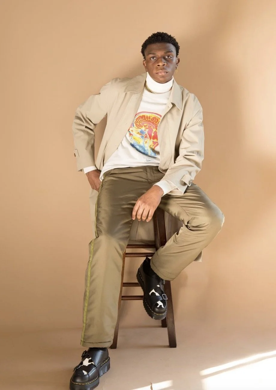 A young man with dark skin sitting on a wooden stool against a beige background. He is wearing a beige trench coat over a white graphic t-shirt with colorful art, khaki pants, and black-and-white chunky shoes. He has short curly hair, earrings, and a gold chain necklace.
