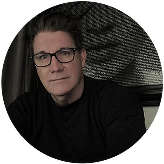 A man with glasses and dark hair, wearing a black shirt, sitting indoors with a window behind him.