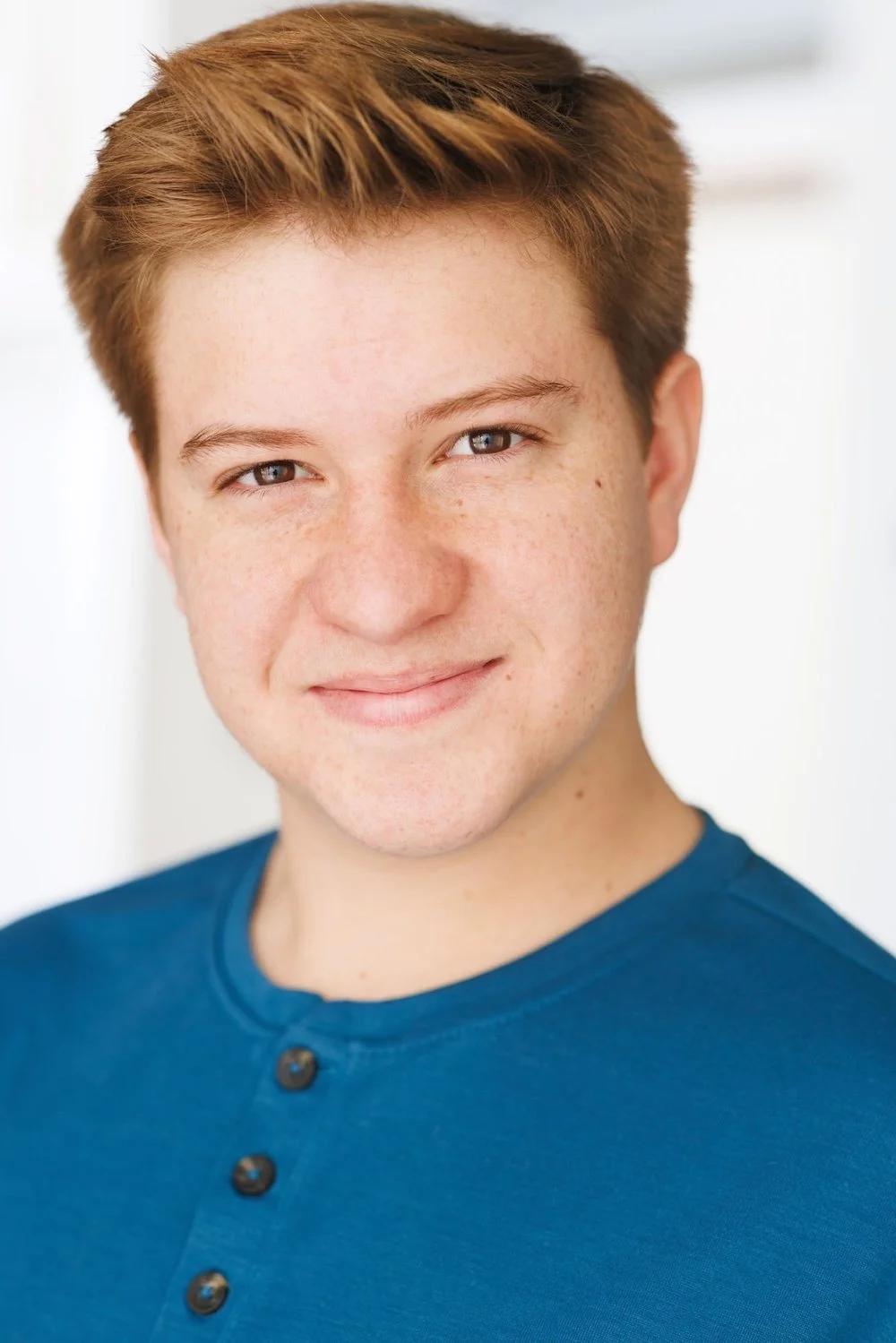 Close-up portrait of a young person with short, red hair and light skin, smiling gently, wearing a blue shirt.