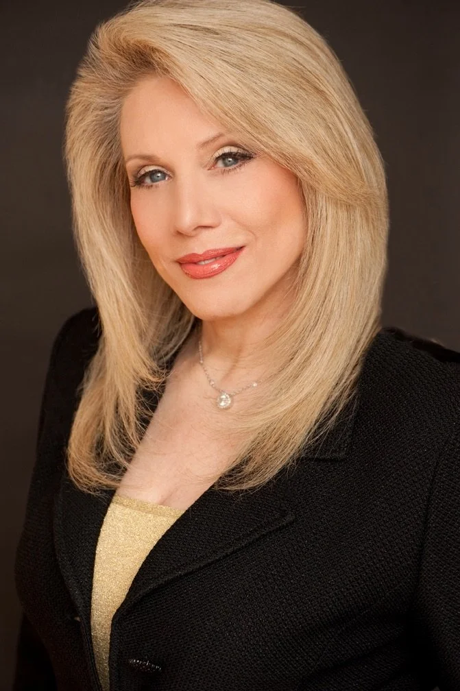 A portrait of a blonde woman smiling, wearing a black blazer and a gold top, with a necklace, across a dark background.