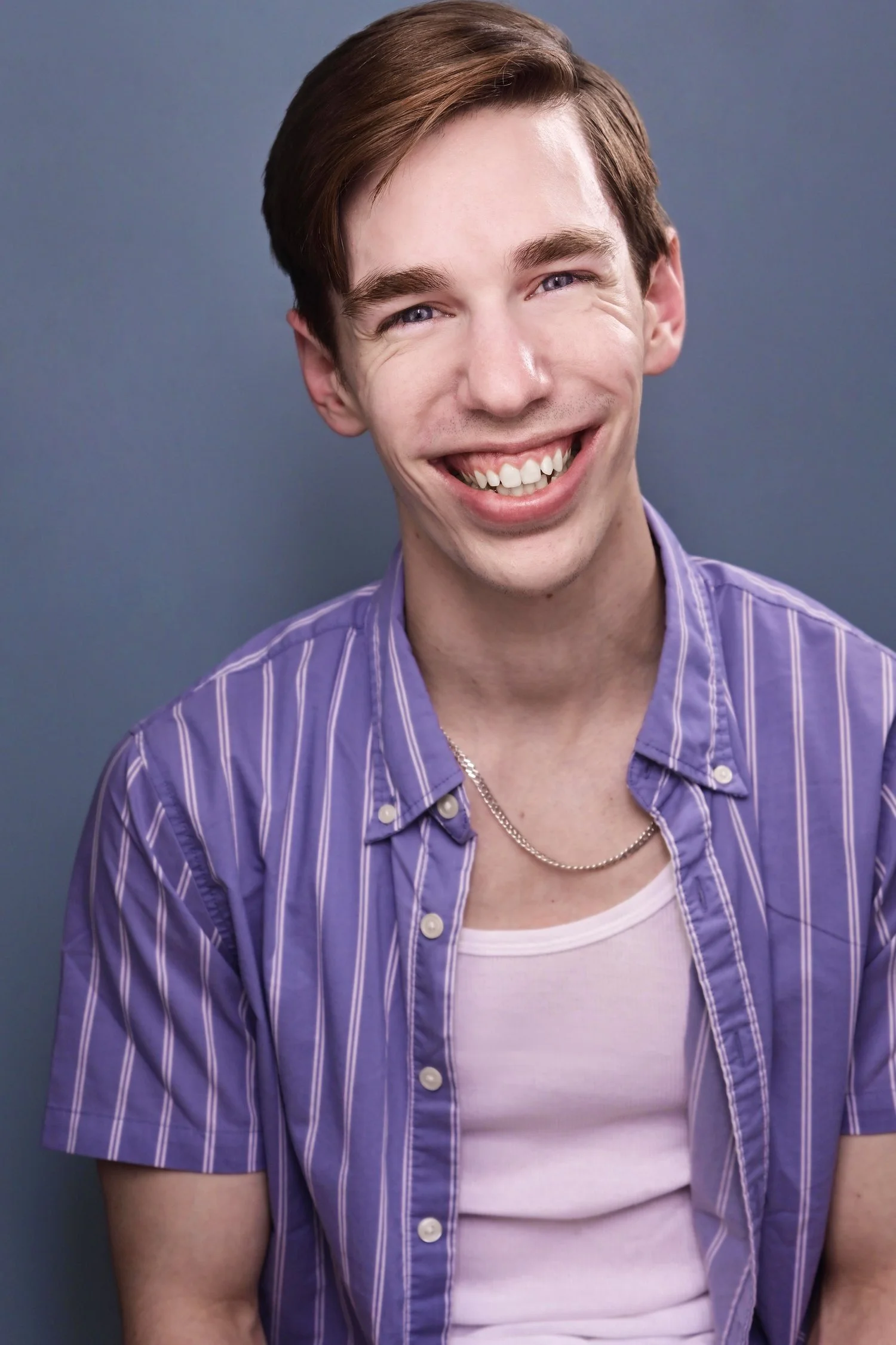 Smiling young man with short brown hair, wearing a purple striped shirt over a white tank top and a silver chain necklace, against a plain gray background.