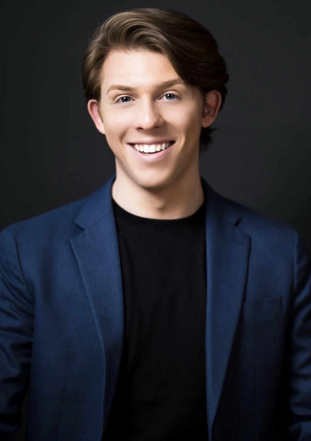 A young man with light brown hair, blue eyes, and a wide smile, dressed in a dark blazer and black shirt, smiling at the camera against a dark background.