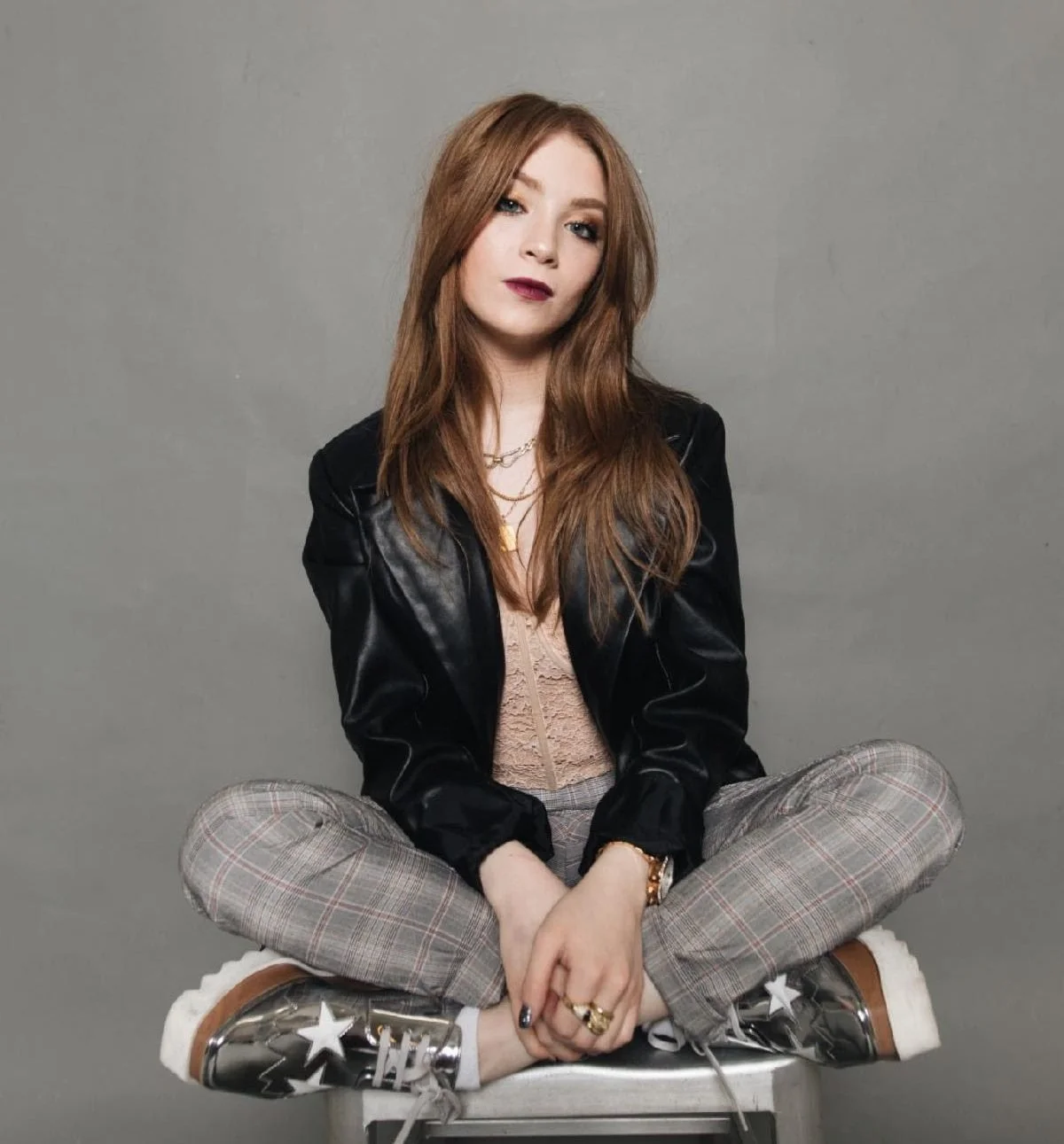 A young woman with long red hair sitting cross-legged on a stool against a plain gray background, wearing a black leather jacket, plaid pants, and shiny high-top sneakers.