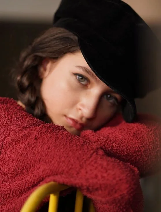 A young woman with short brown hair, blue eyes, and light skin, wearing a black hat and a red textured sweater, leaning on a yellow chair, resting her head on her arms, looking at the camera with a soft expression.