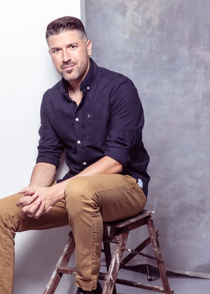 A man with short dark hair and a trimmed beard, wearing a navy blue button-up shirt and tan pants, sitting on a small wooden stool against a plain background.