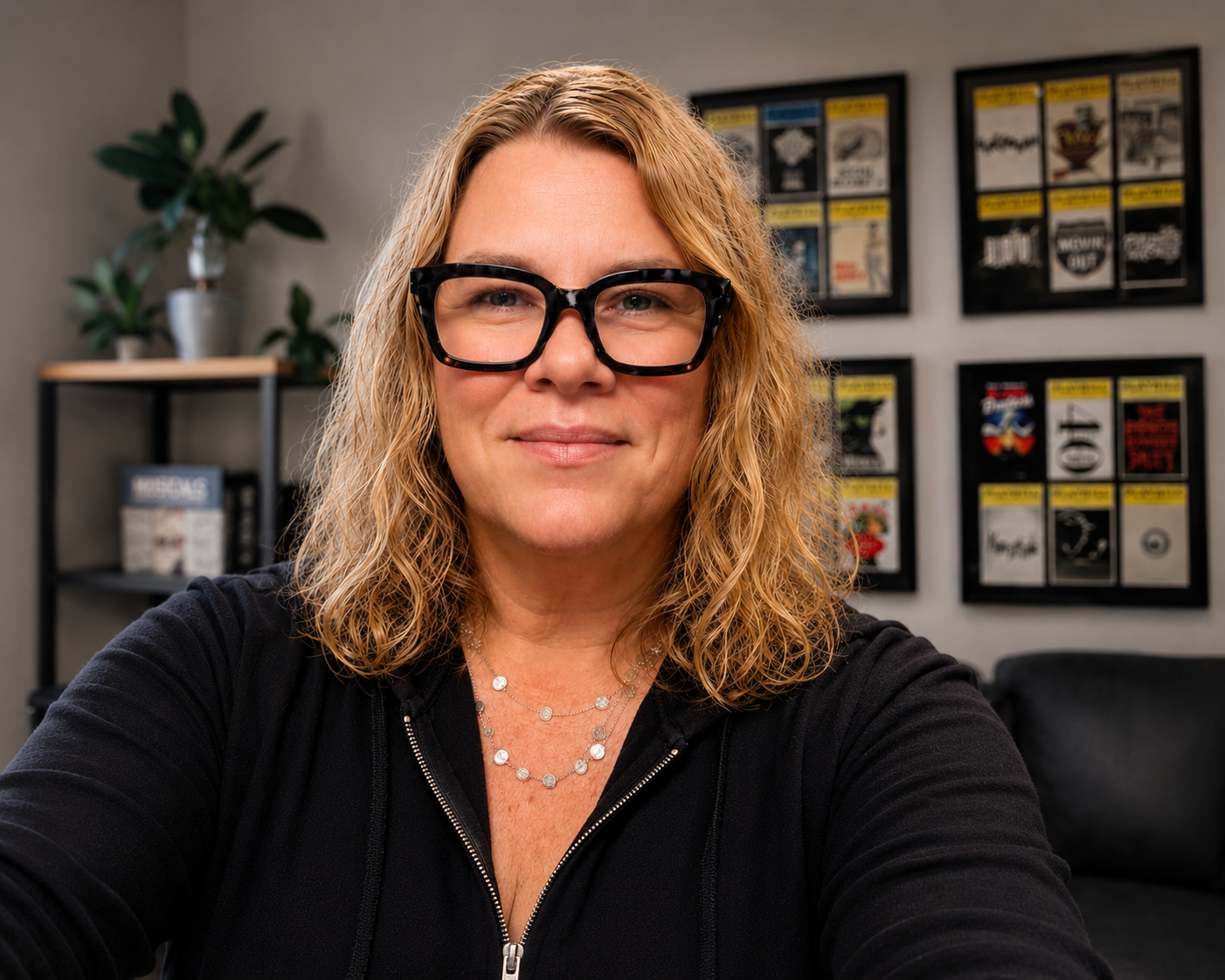 A smiling woman with curly blonde hair, wearing black glasses, a black zip-up jacket, and layered necklaces, sitting in an office with framed magazines on the wall and plants on a shelf.
