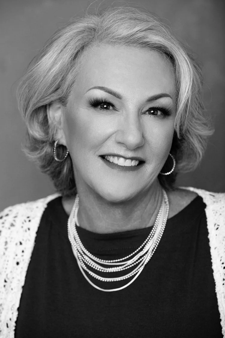 Black and white portrait of a smiling middle-aged woman with short, wavy gray hair, wearing hoop earrings, layered pearl necklaces, and a black top with a white crochet cardigan.