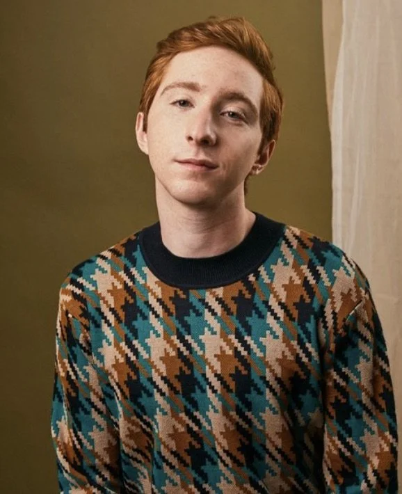 Portrait of a Larry Saperstein with reddish hair wearing a patterned sweater with brown, black, blue, and beige colors, standing in front of a plain background.