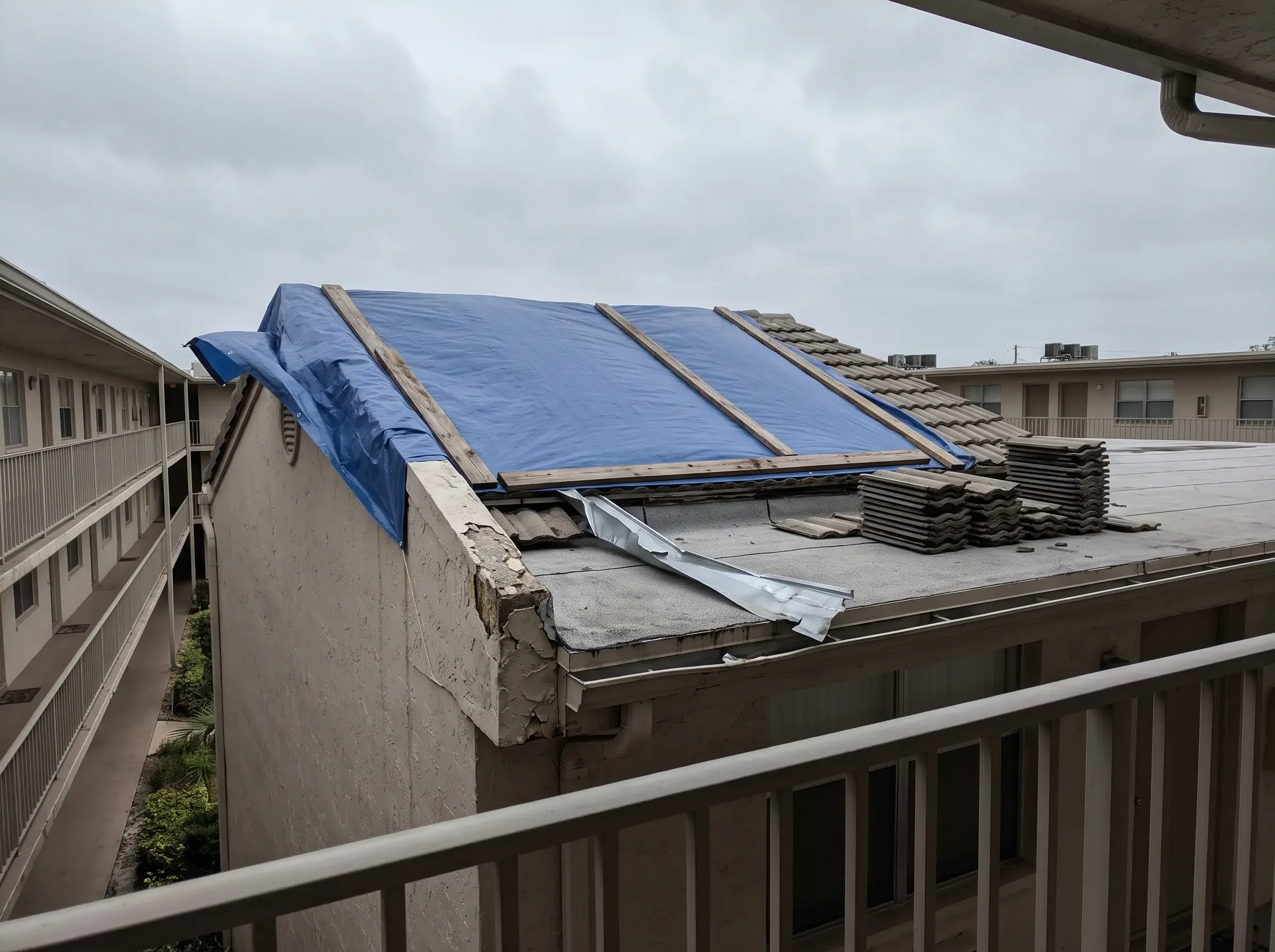 A tarped section of a condominium building roof seen from an upper walkway, with damaged parapet tiles stacked below