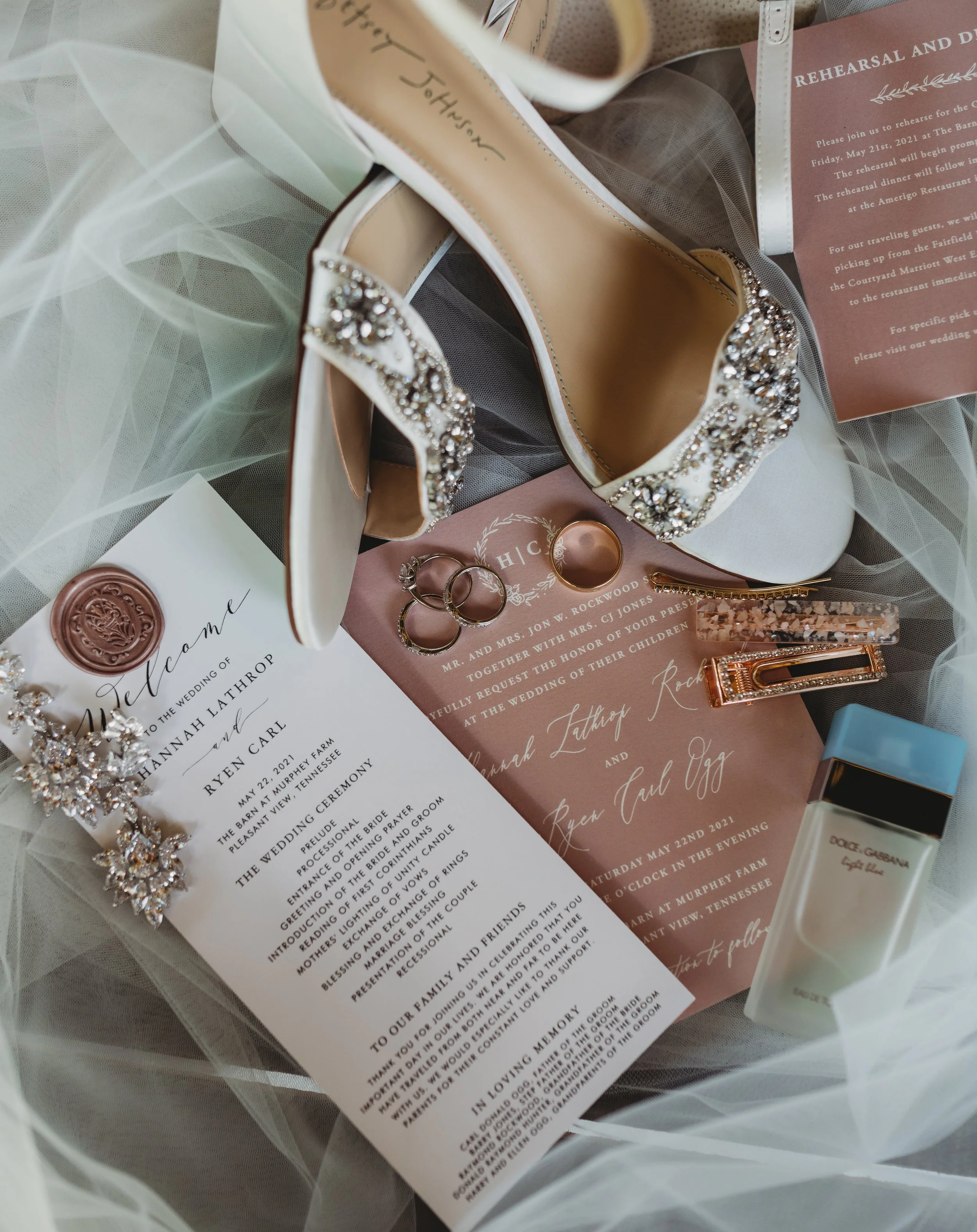 Wedding invitation, a pair of embellished high heel shoes, rings, and perfume bottle on tulle fabric, wedding rehearsal and ceremony details.