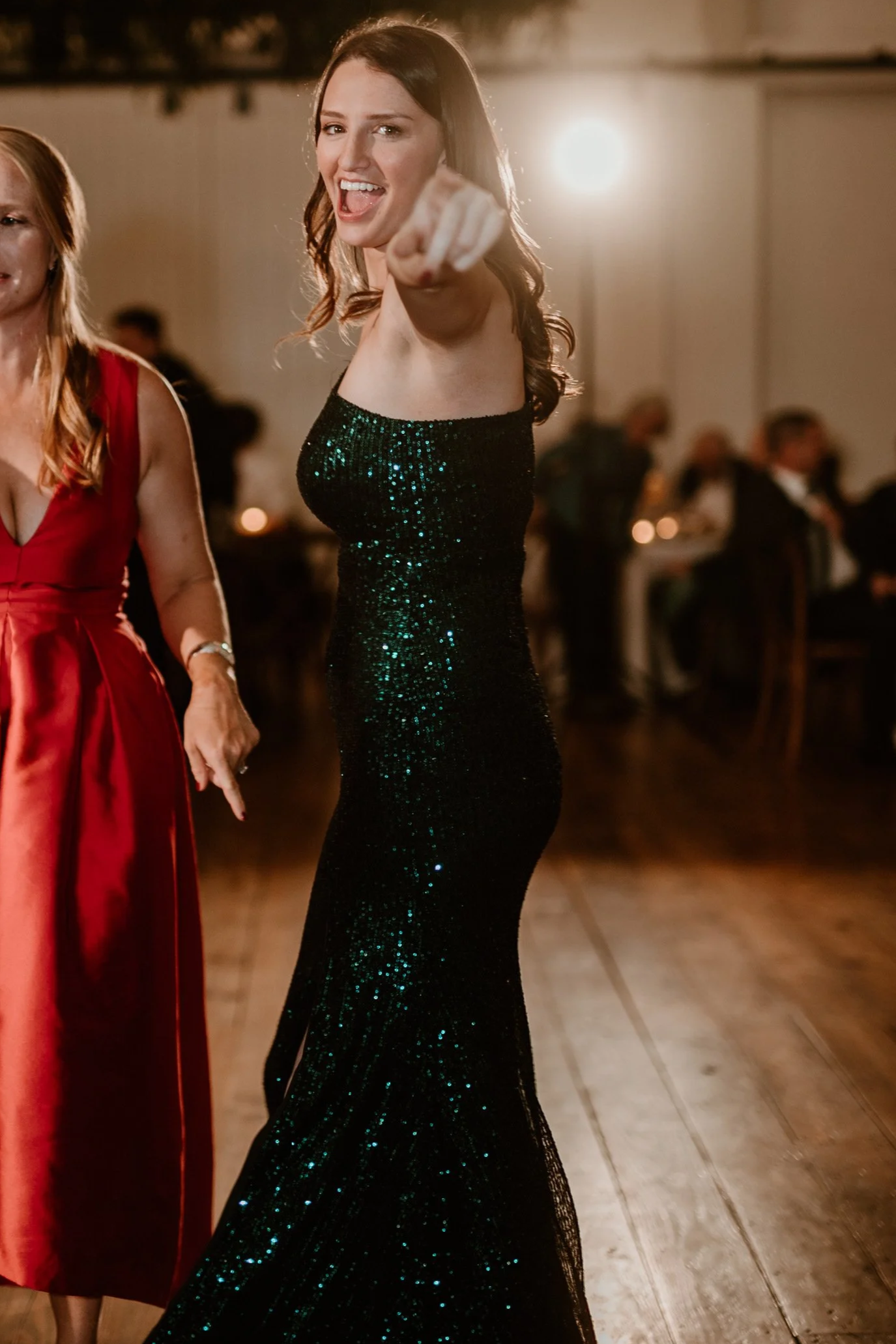 A woman in a black, glittery, sleeveless dress is smiling playfully and pointing towards the camera at a party or celebration event.