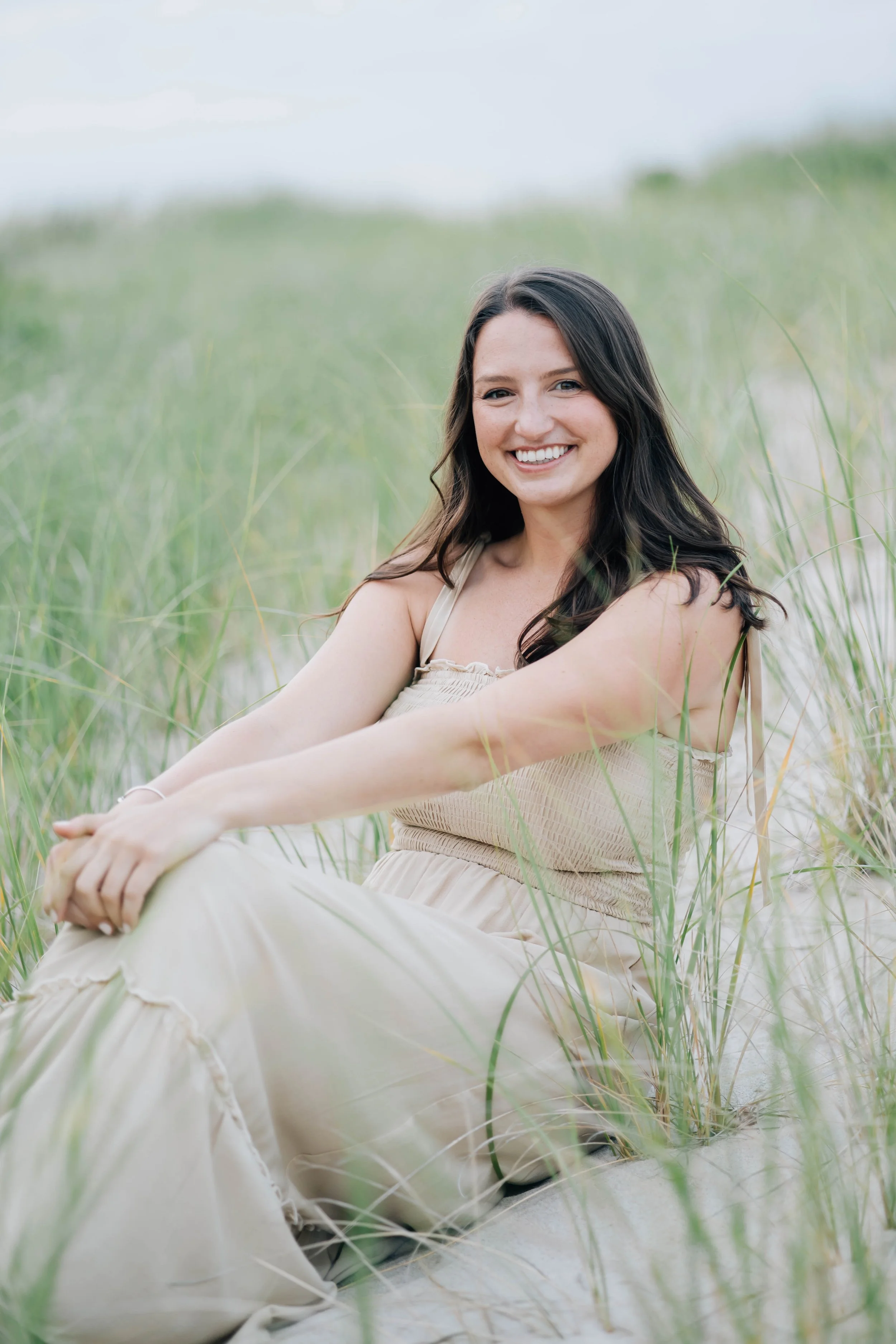 A young woman with long dark hair, smiling, sitting on sand surrounded by tall green grass at the beach on a cloudy day.