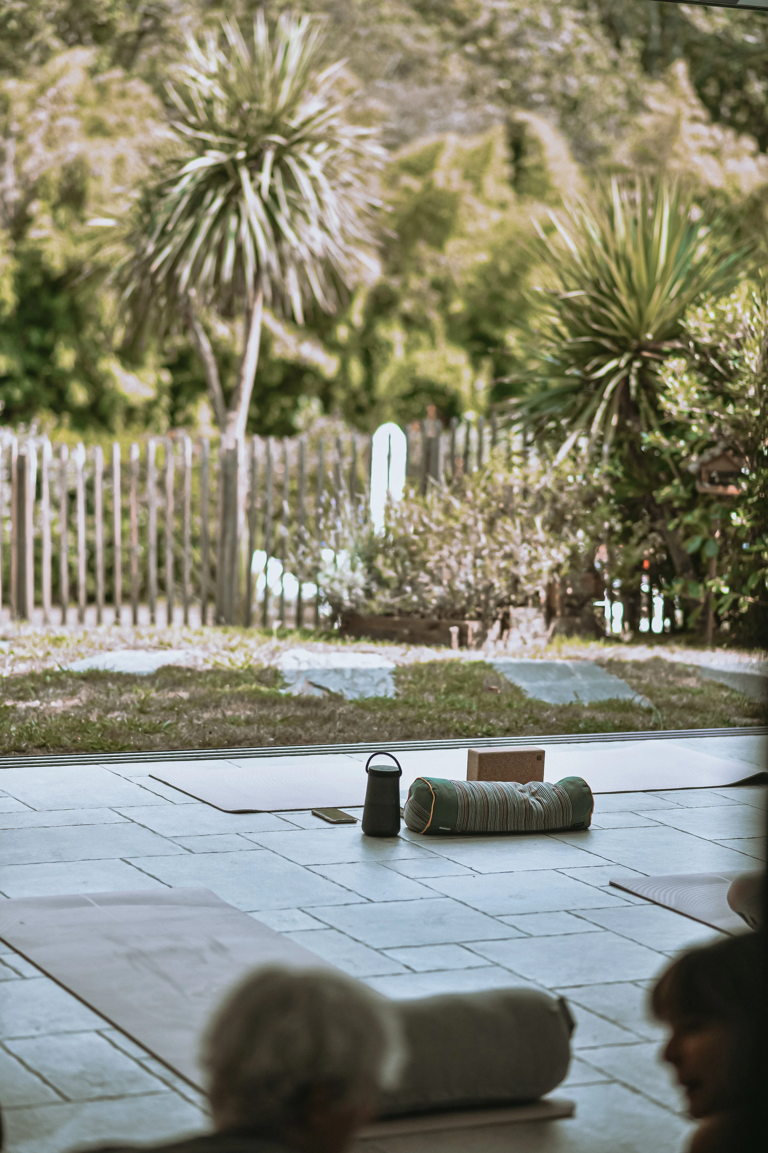 Salle de yoga avec tapis, bouteille d'eau, boîte en bois, et coussin, vue sur un jardin avec des arbres et une clôture.