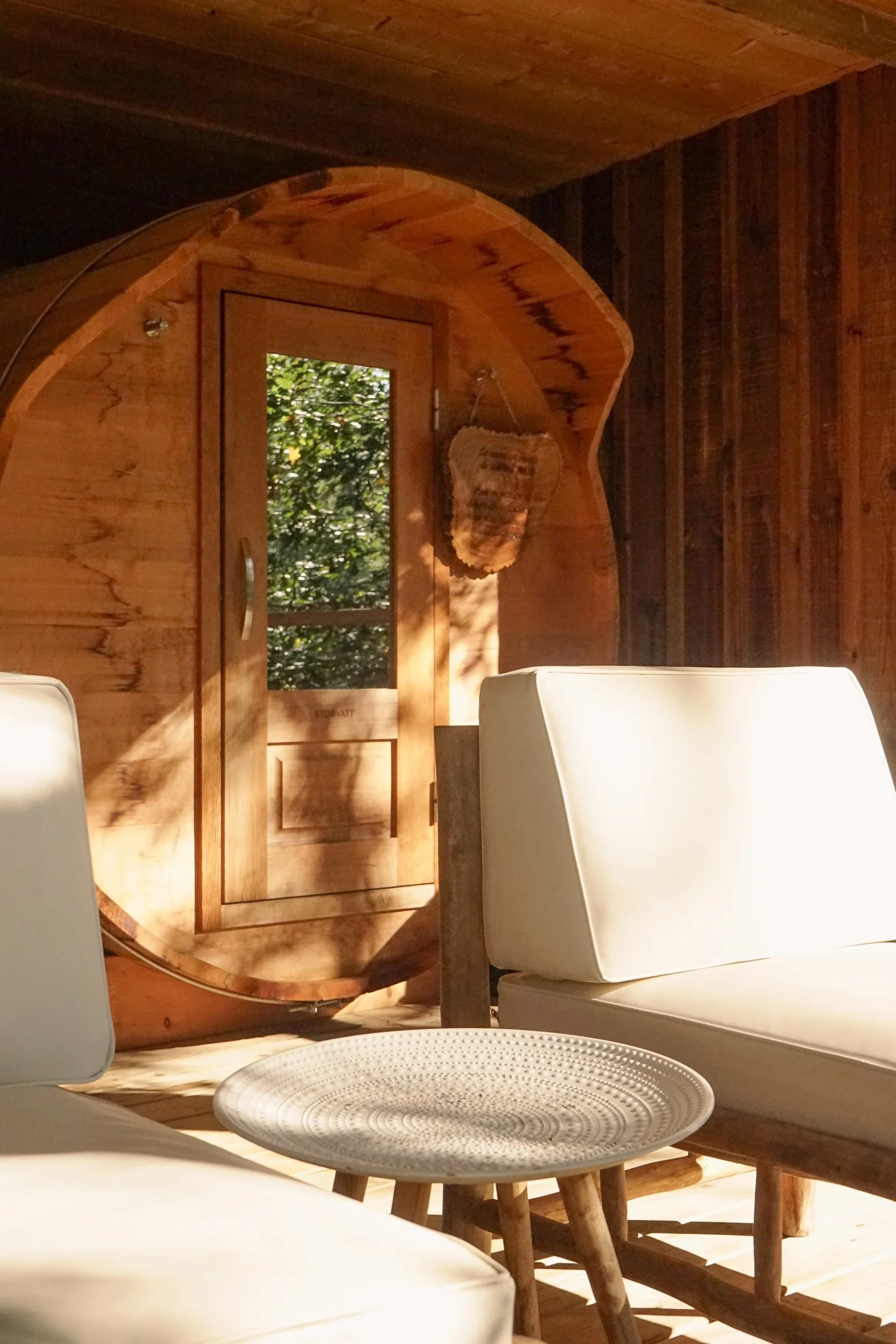 Intérieur d'une pièce en bois avec un miroir rond, un siège blanc, une table en bois, et une porte en bois avec une vue sur la nature extérieure.