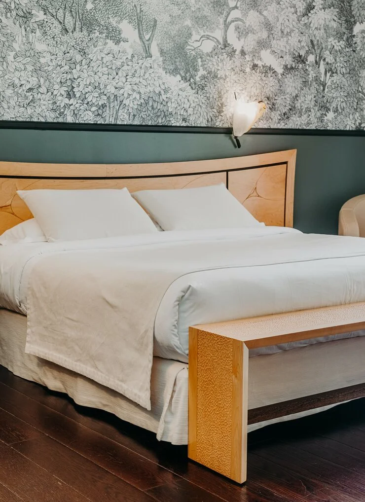 Lits dans une chambre avec une tête de lit en bois clair et une nappe blanche, mural avec une fresque de nature monochrome, luminaire mural en forme d'oiseau, sol en bois foncé.