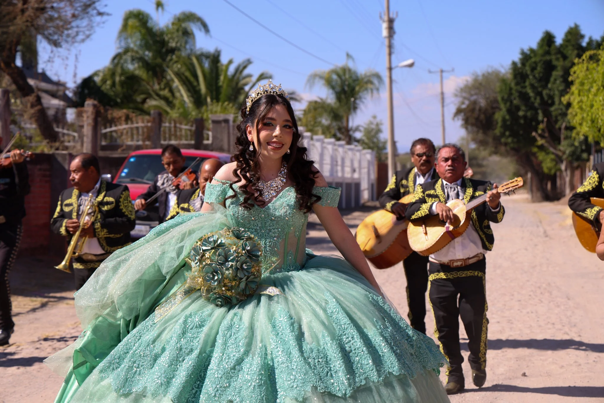 A young woman in a mint green quinceañera dress with lace and floral details, wearing a tiara and jewelry, smiling while walking outdoors during a celebration with musicians in traditional Mexican attire playing guitars and violins in the background.