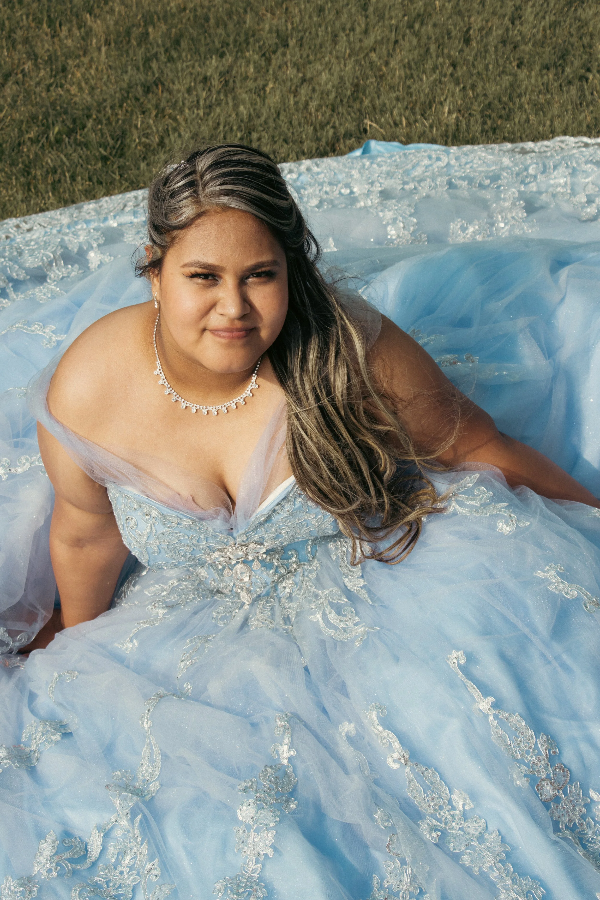 A woman in a light blue, embroidered ball gown lying on the grass.